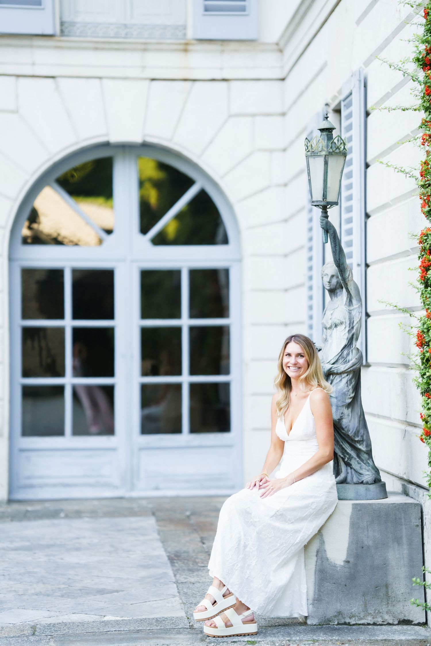 Person in a flowing white dress sits elegantly on stone steps by an arched window and vintage lamp post.