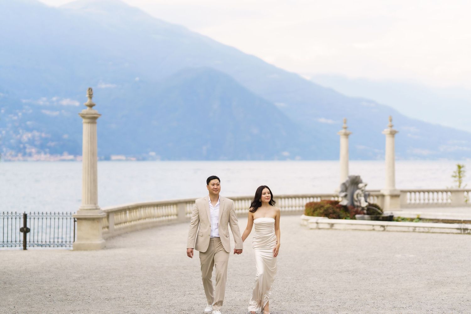 A couple walks hand in hand along a scenic stone balcony overlooking a coastal landscape on a bright sunny day.