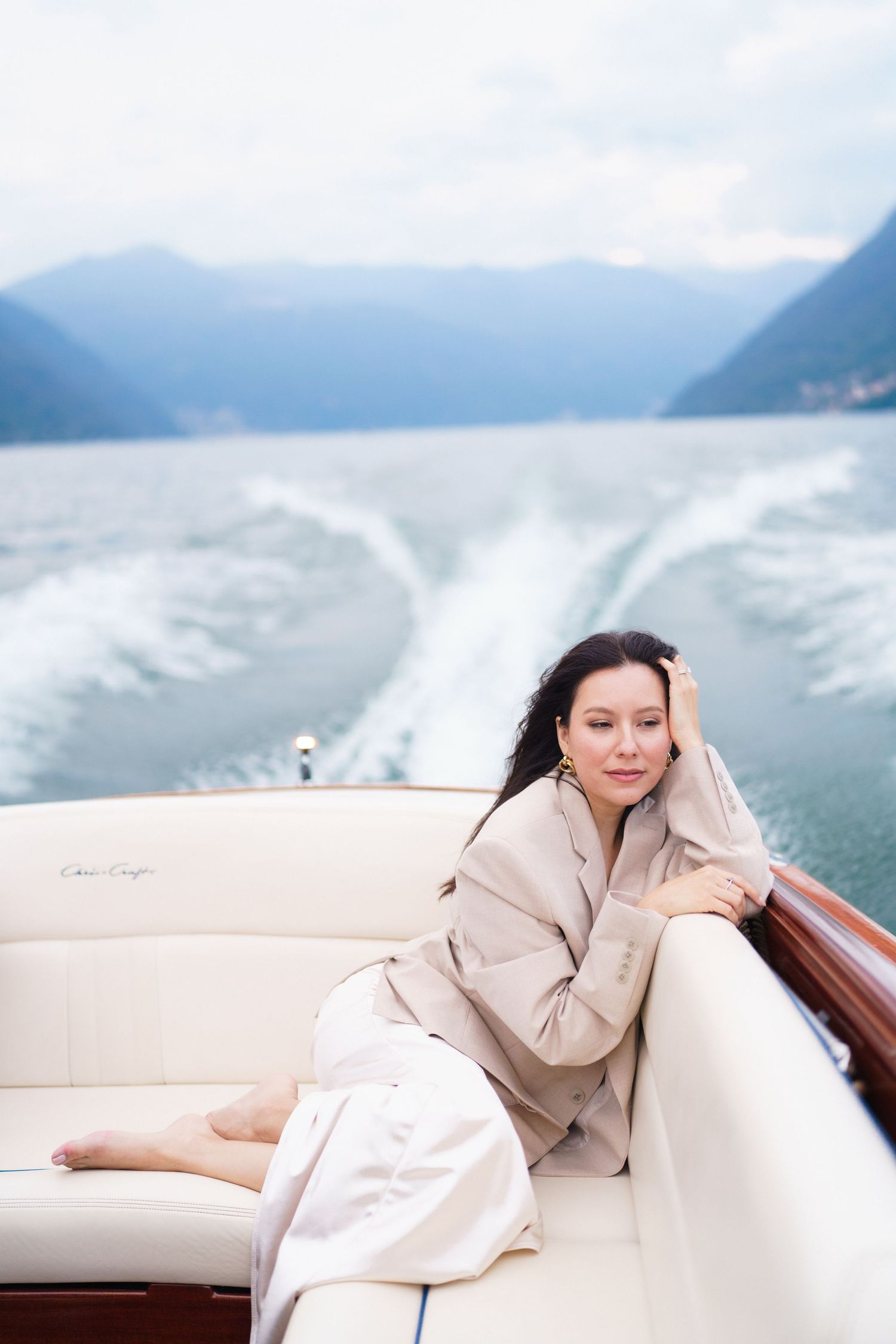 Person lounges on white boat cushions wearing beige coat while cruising past misty mountains on a lake.