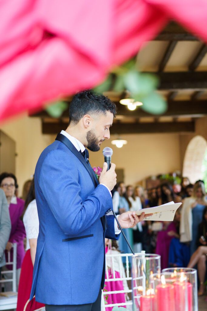 Person in blue suit speaks into microphone during wedding ceremony with pink decor.