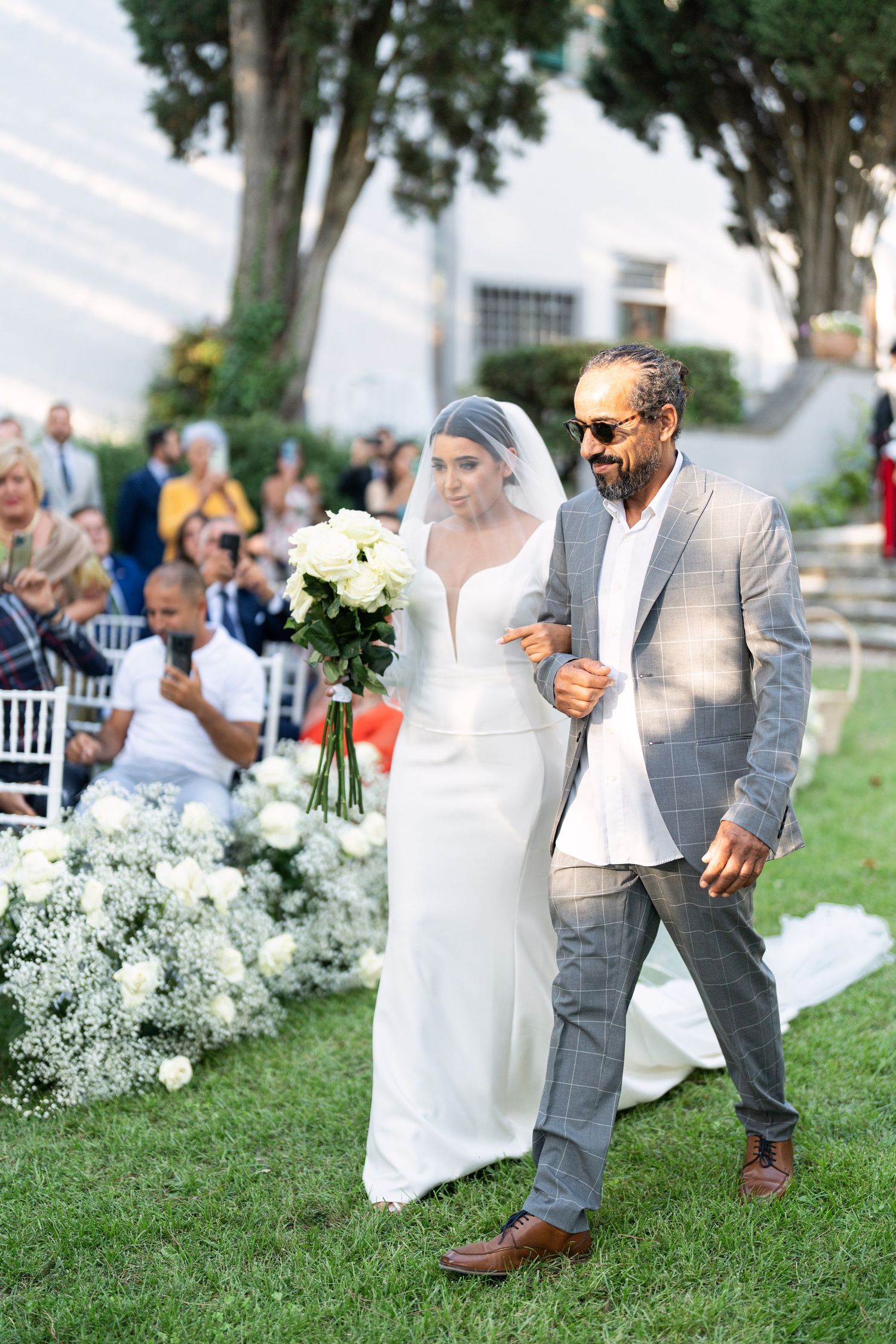 Wedding couple strolls on grassy lawn with white flowers and outdoor seating in background.
