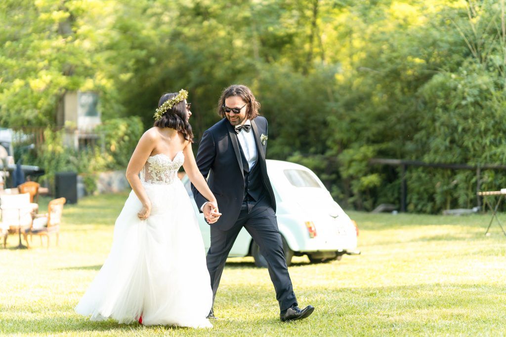 Wedding celebration on a grassy lawn with guests walking near a contemporary glass structure.