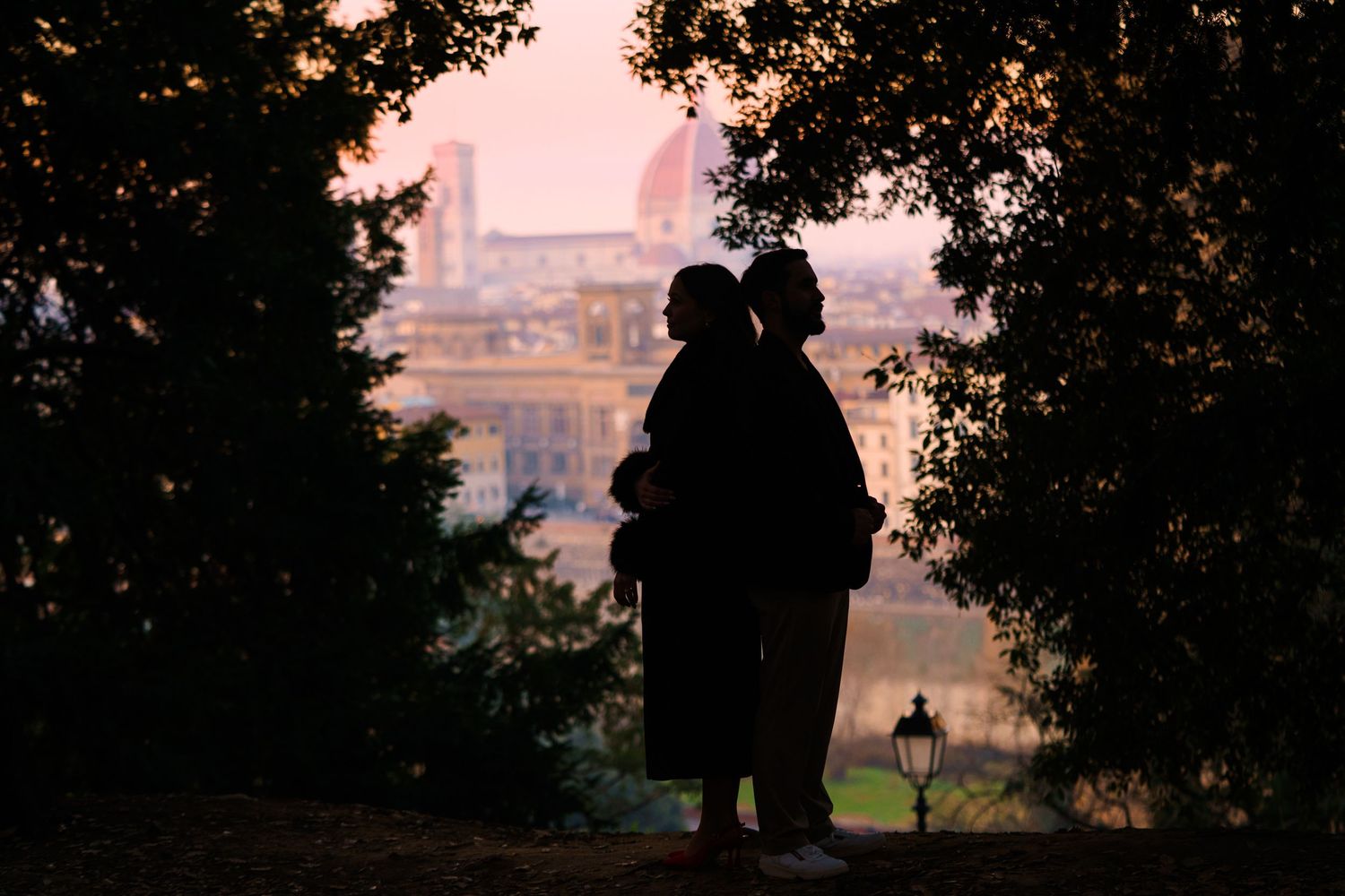 Silhouettes of two people standing together at sunset with a distant cityscape and church dome visible through trees.