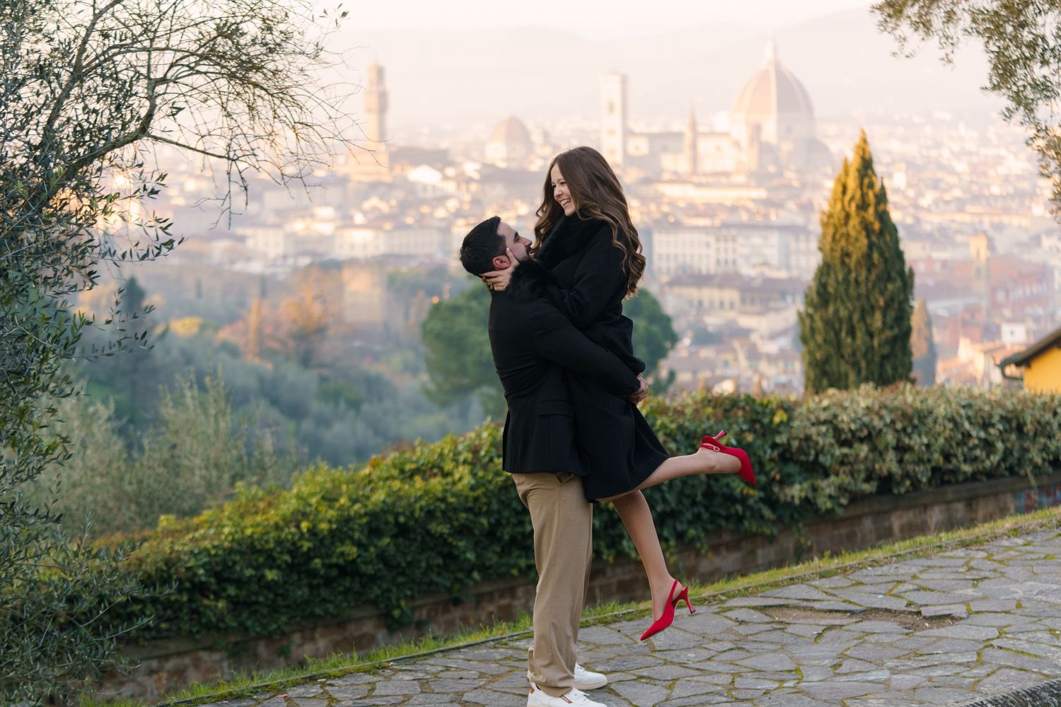 A couple embraces on a scenic overlook with a cityscape visible in the background during sunset.