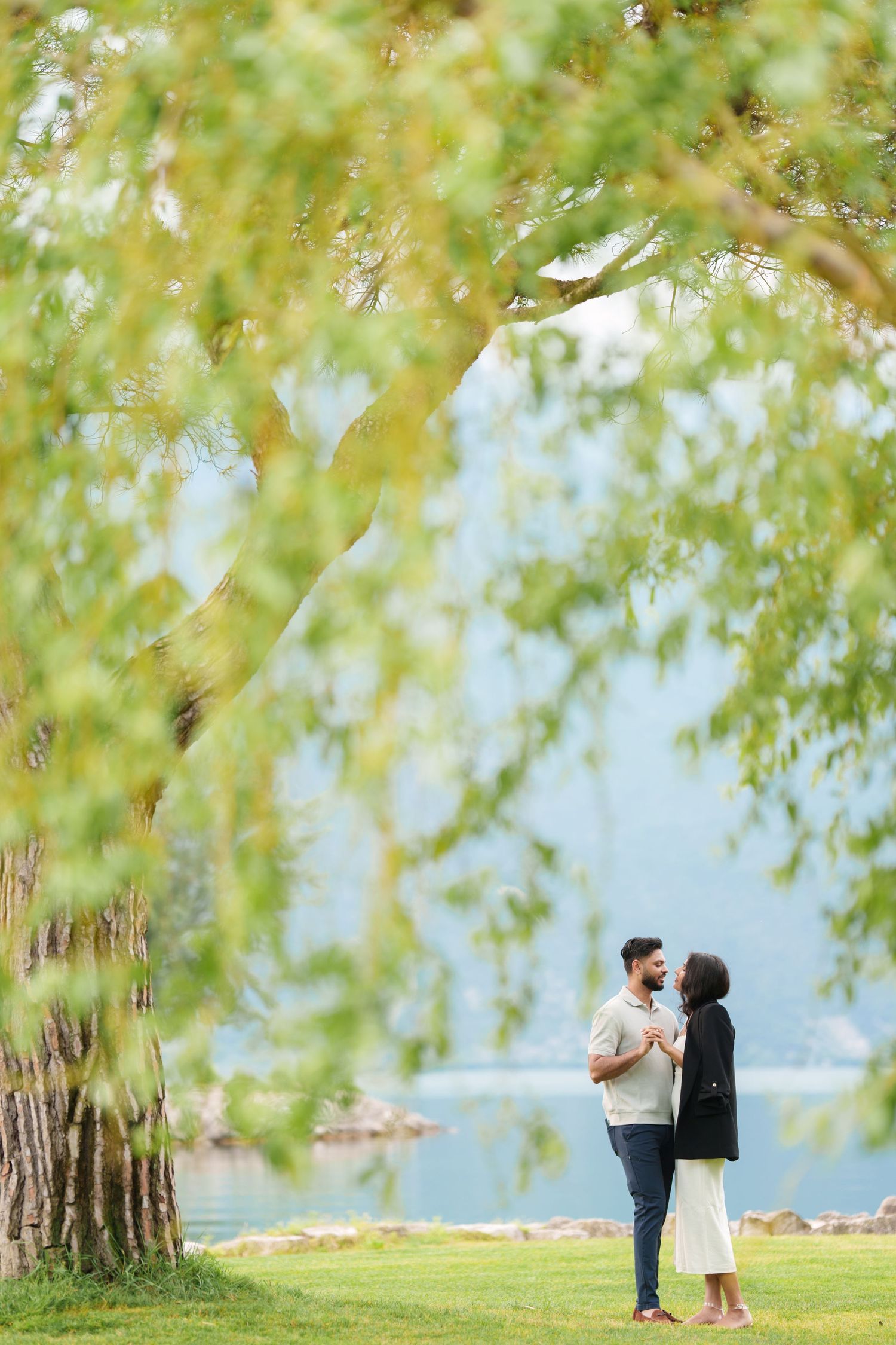 A couple shares a romantic moment under weeping willow trees by a tranquil lakeside on a sunny day.