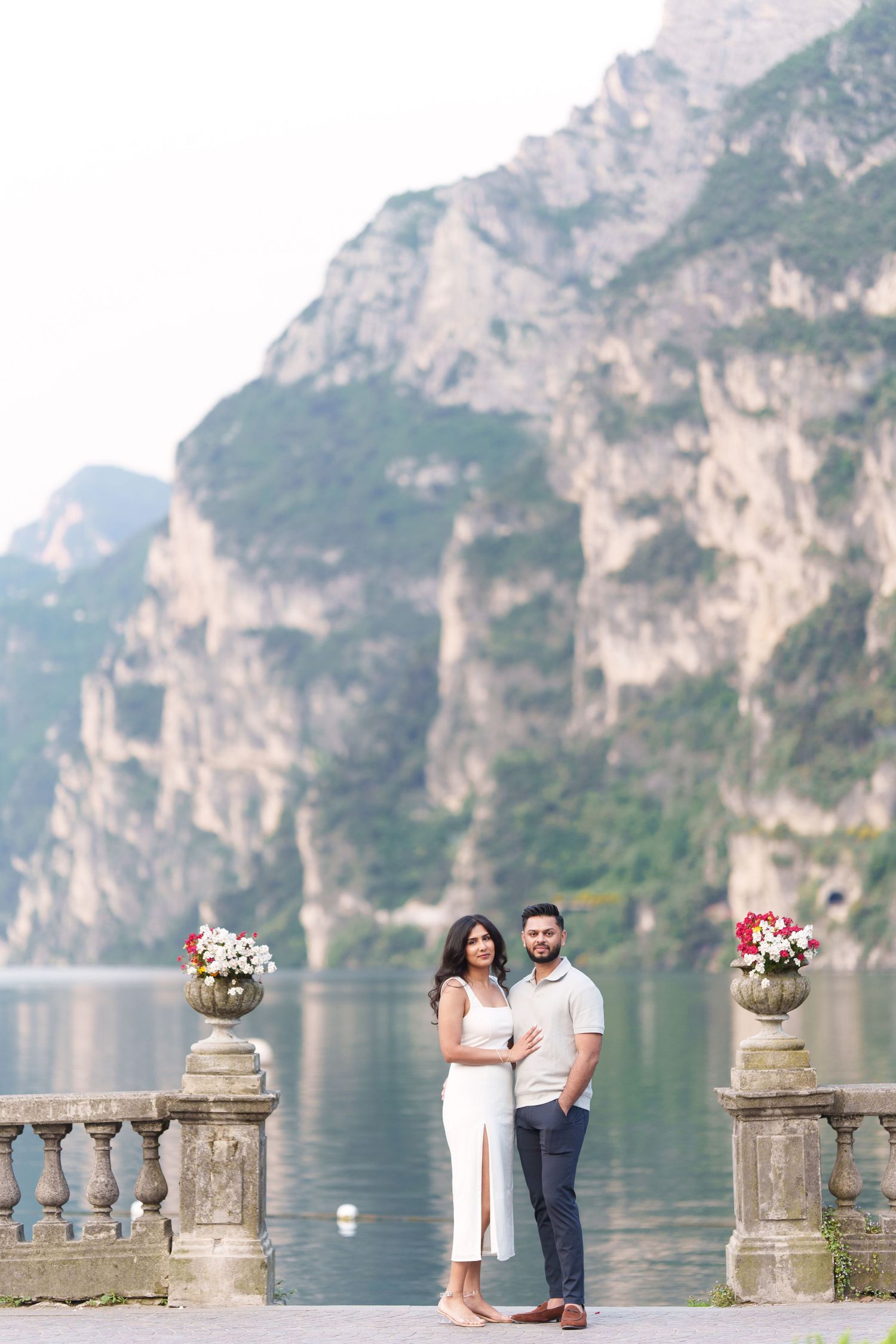Couple in white outfits standing on stone balcony overlooking scenic Lake Como in Italy with mountains in background.