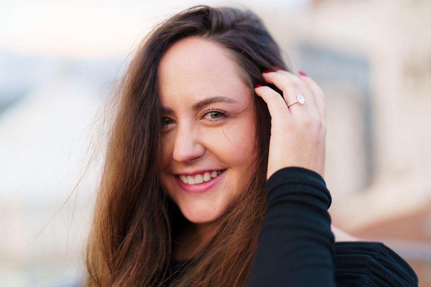A person with long brown hair smiles warmly while standing outdoors in natural lighting.
