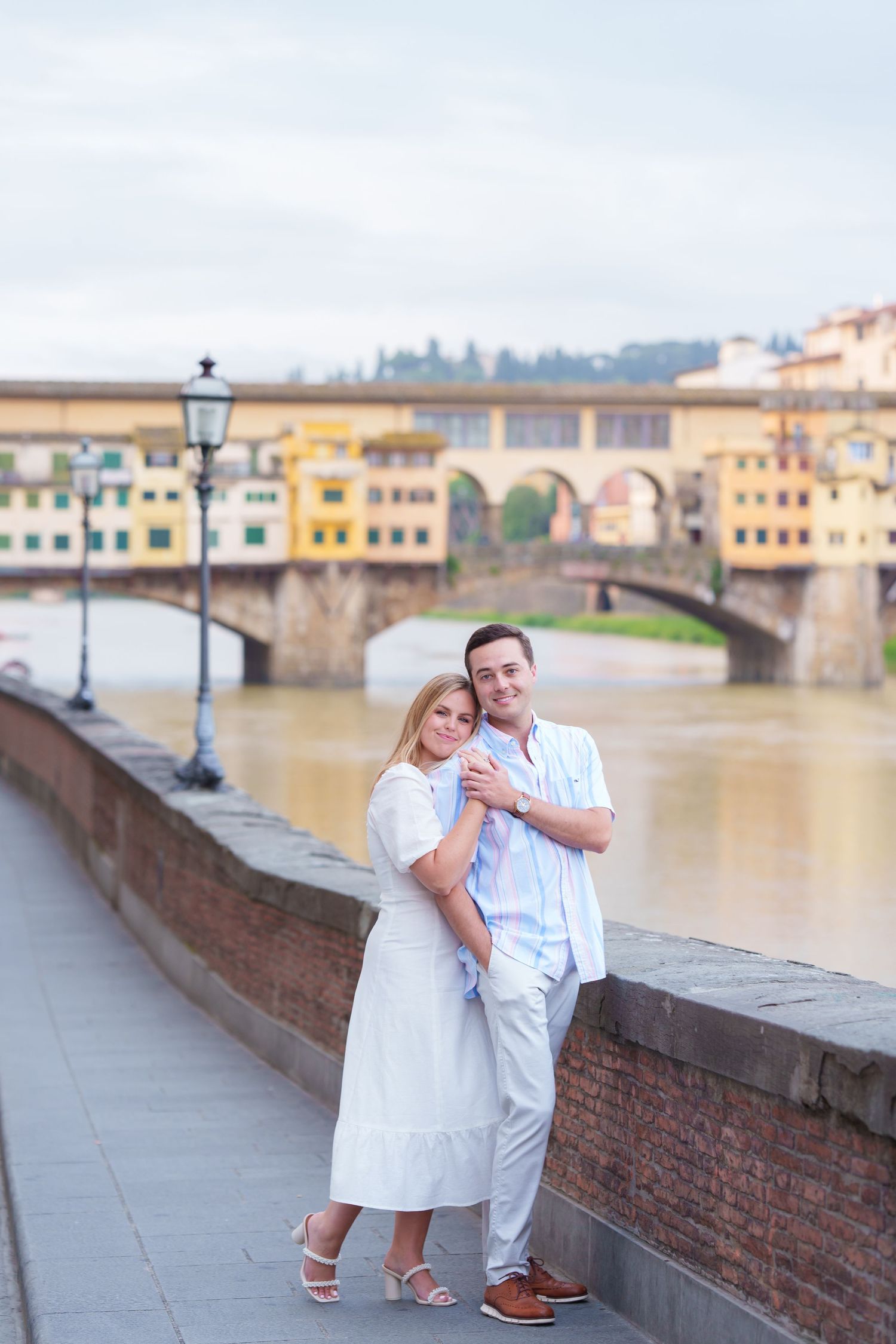 A couple in white outfits poses along a riverbank in Florence, Italy with historic buildings and bridges in the background.