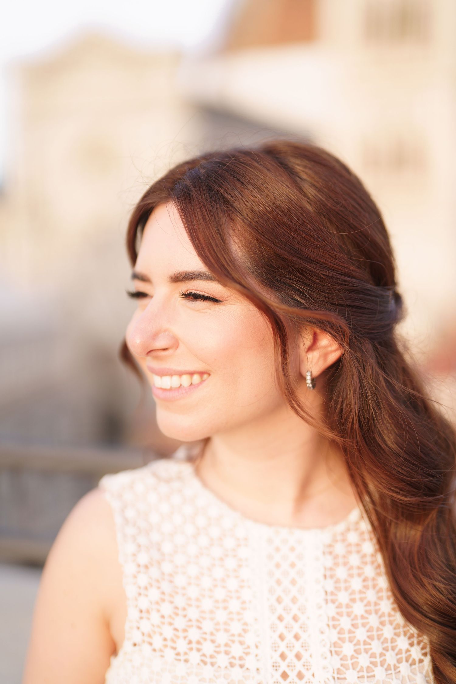 A person with brown hair smiles warmly while wearing a white lace top in soft natural lighting.