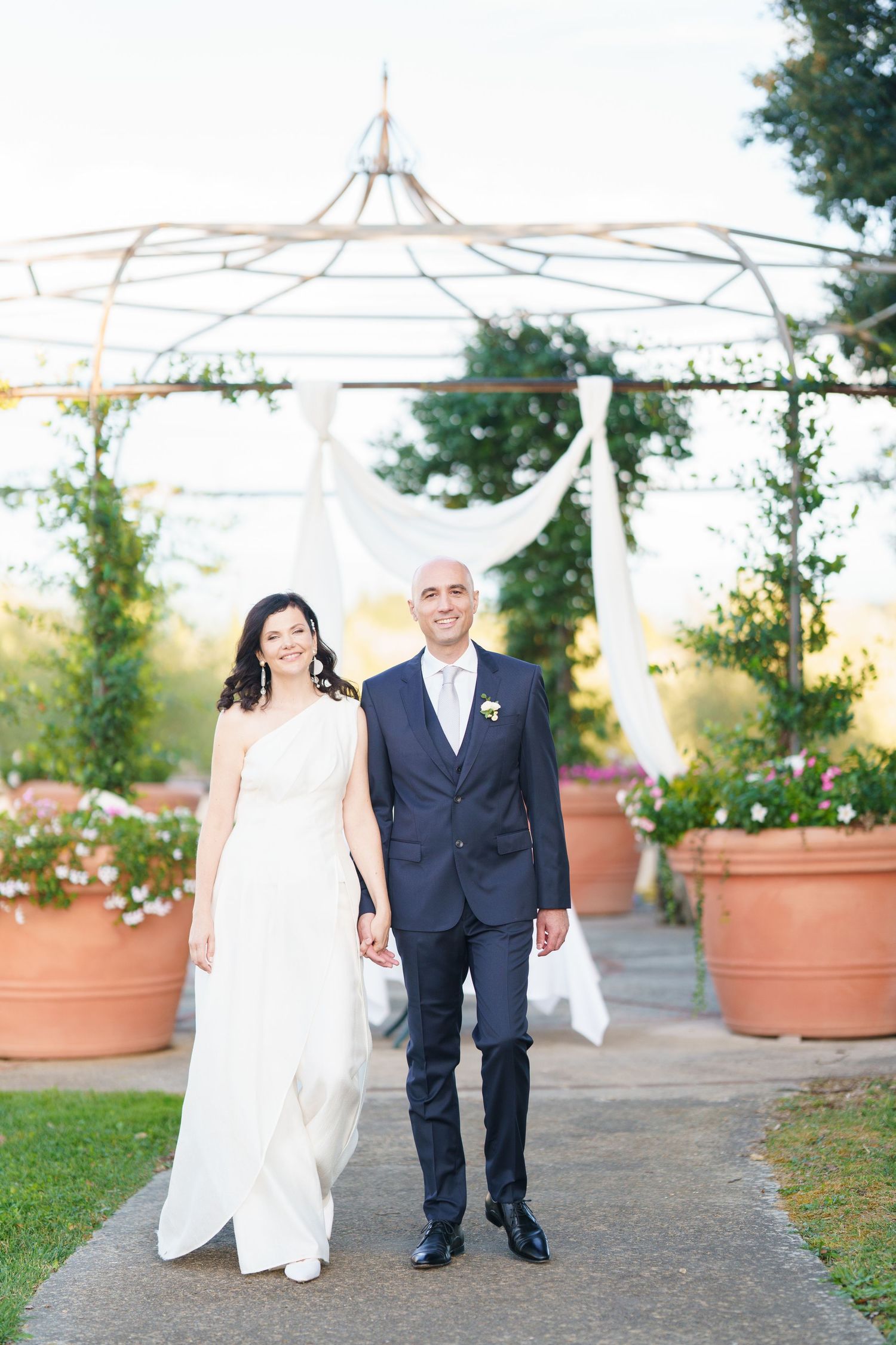 Couple in wedding attire walking together on garden path with white tent structure.