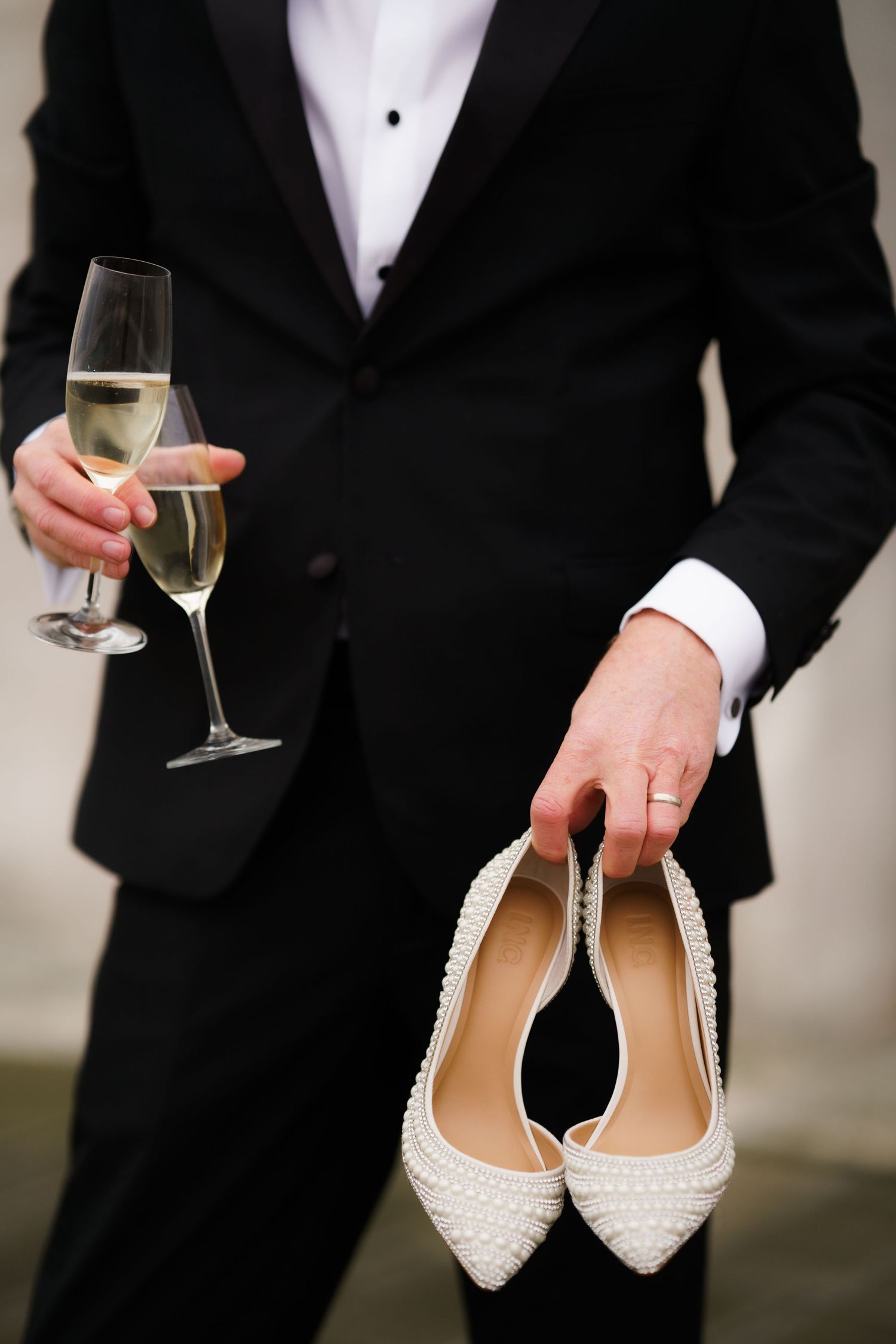 Person in black tuxedo holds champagne glass and white beaded bridal shoes at formal event.