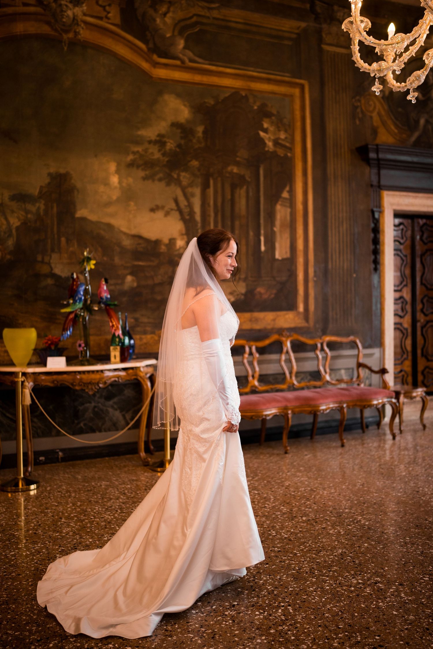 A bride in a flowing white wedding dress poses gracefully in an ornate baroque palace ballroom.