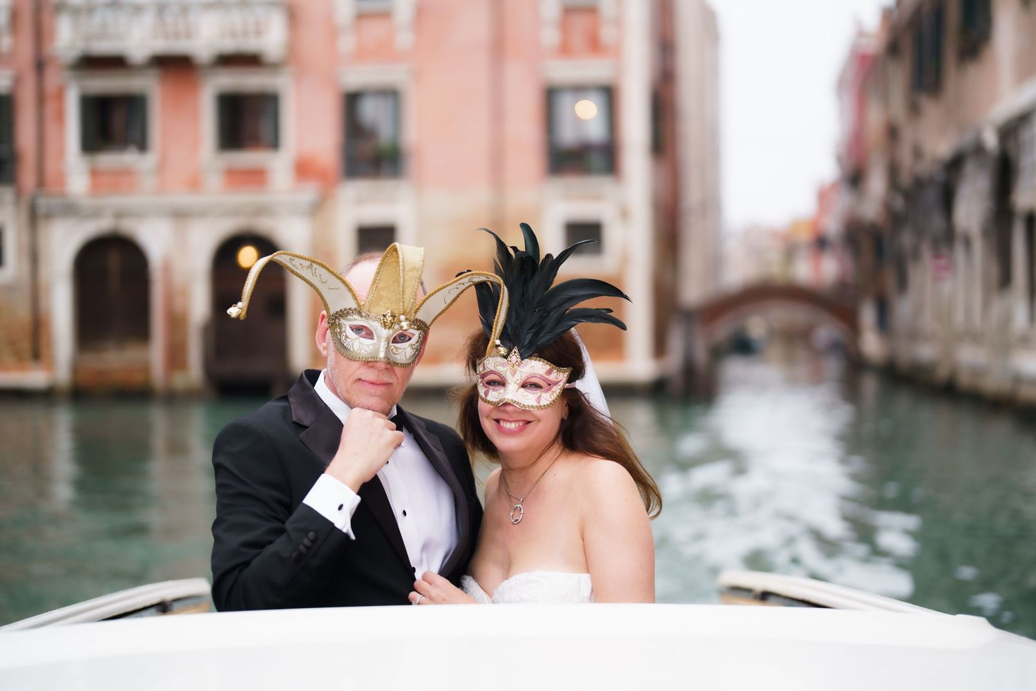 A couple wearing elegant Venetian carnival masks pose together on a gondola in a scenic Venice canal.