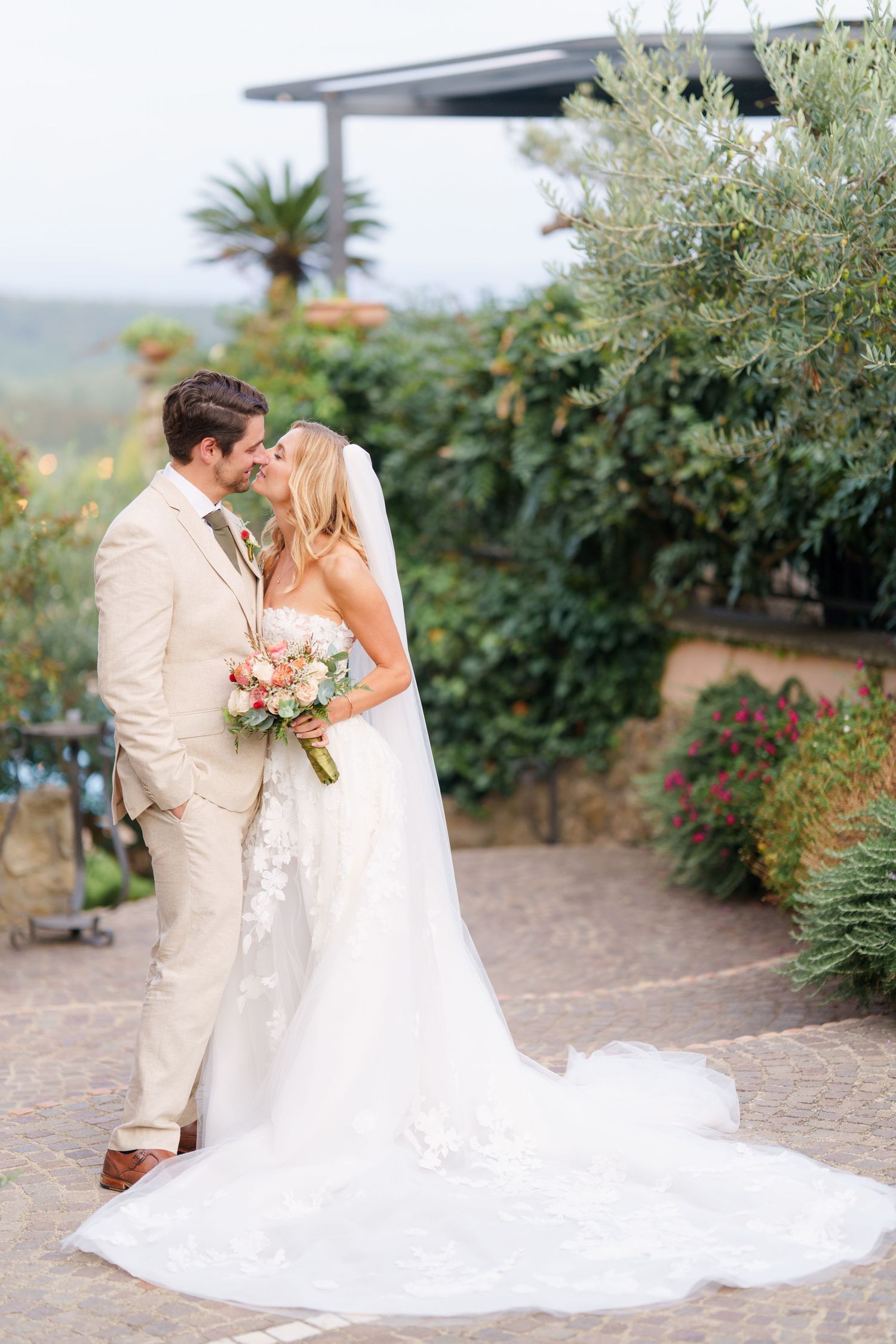 A romantic wedding couple shares an intimate moment in a garden setting with palm trees and lush greenery.