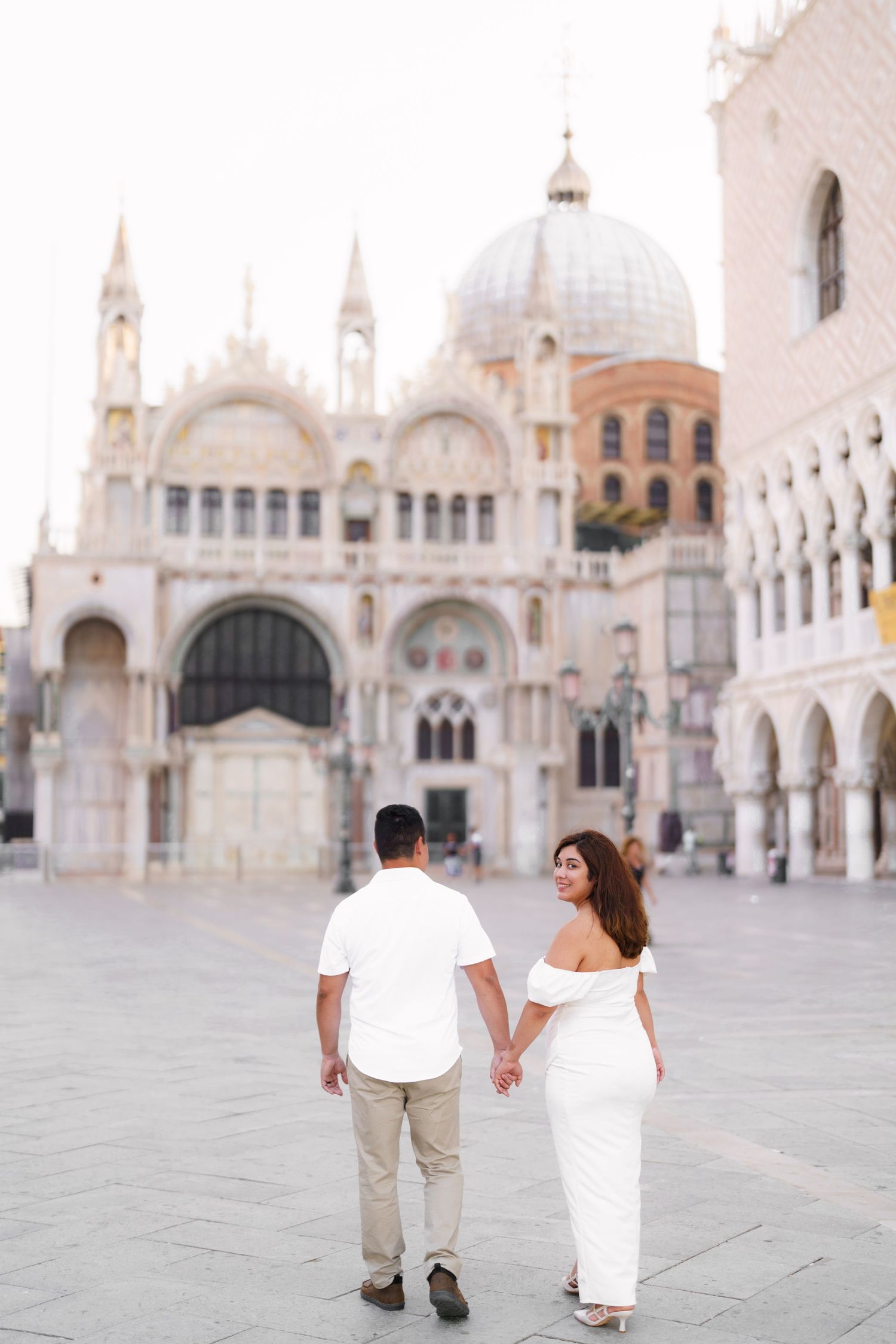 Couple holding hands walking through St. Mark's Square in Venice with the basilica and architecture in the background.