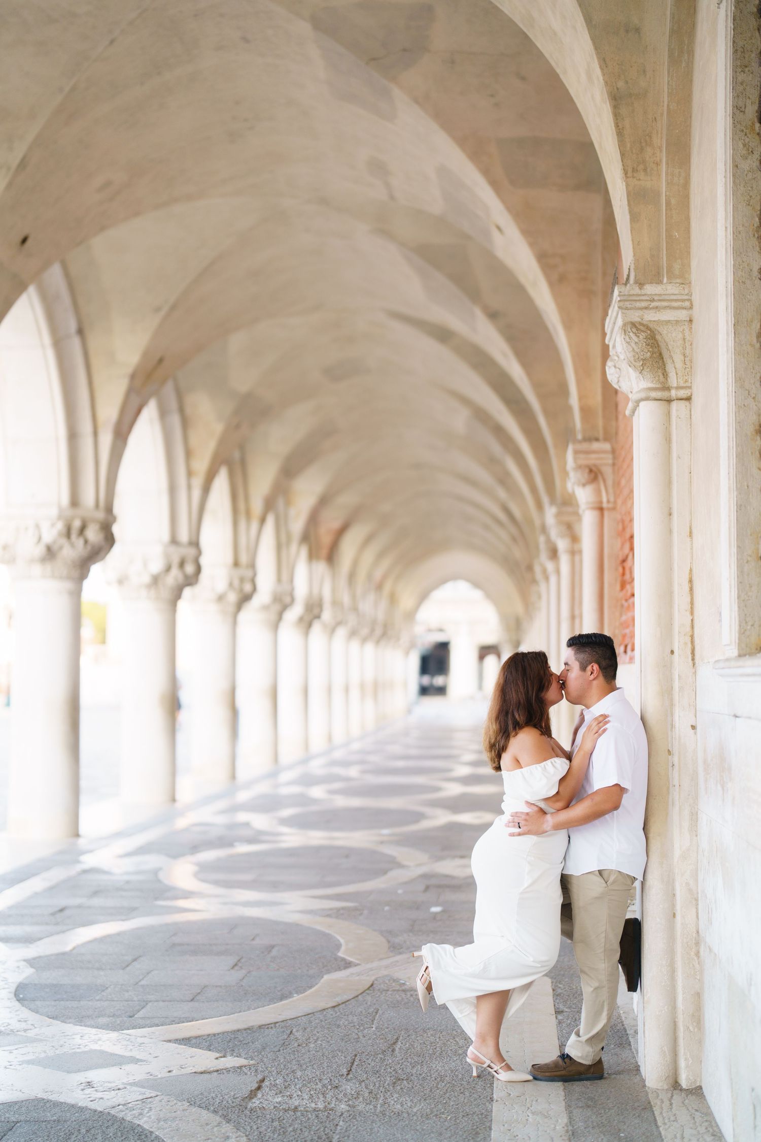 A couple shares an intimate moment in a white arched colonnade walkway with repeating architectural columns.