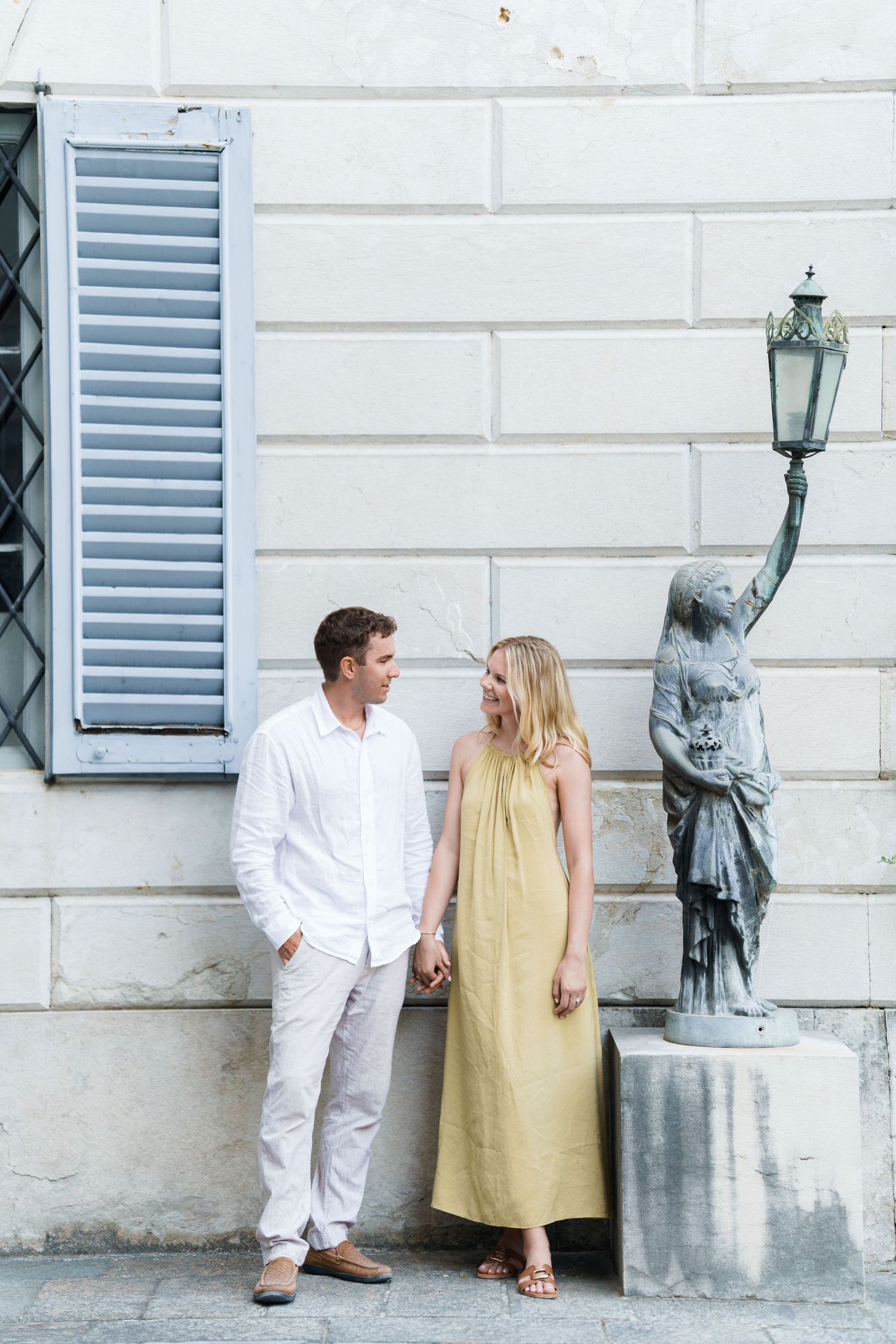 Couple holding hands in white and yellow outfits beside a classical statue and shuttered window in Europe.
