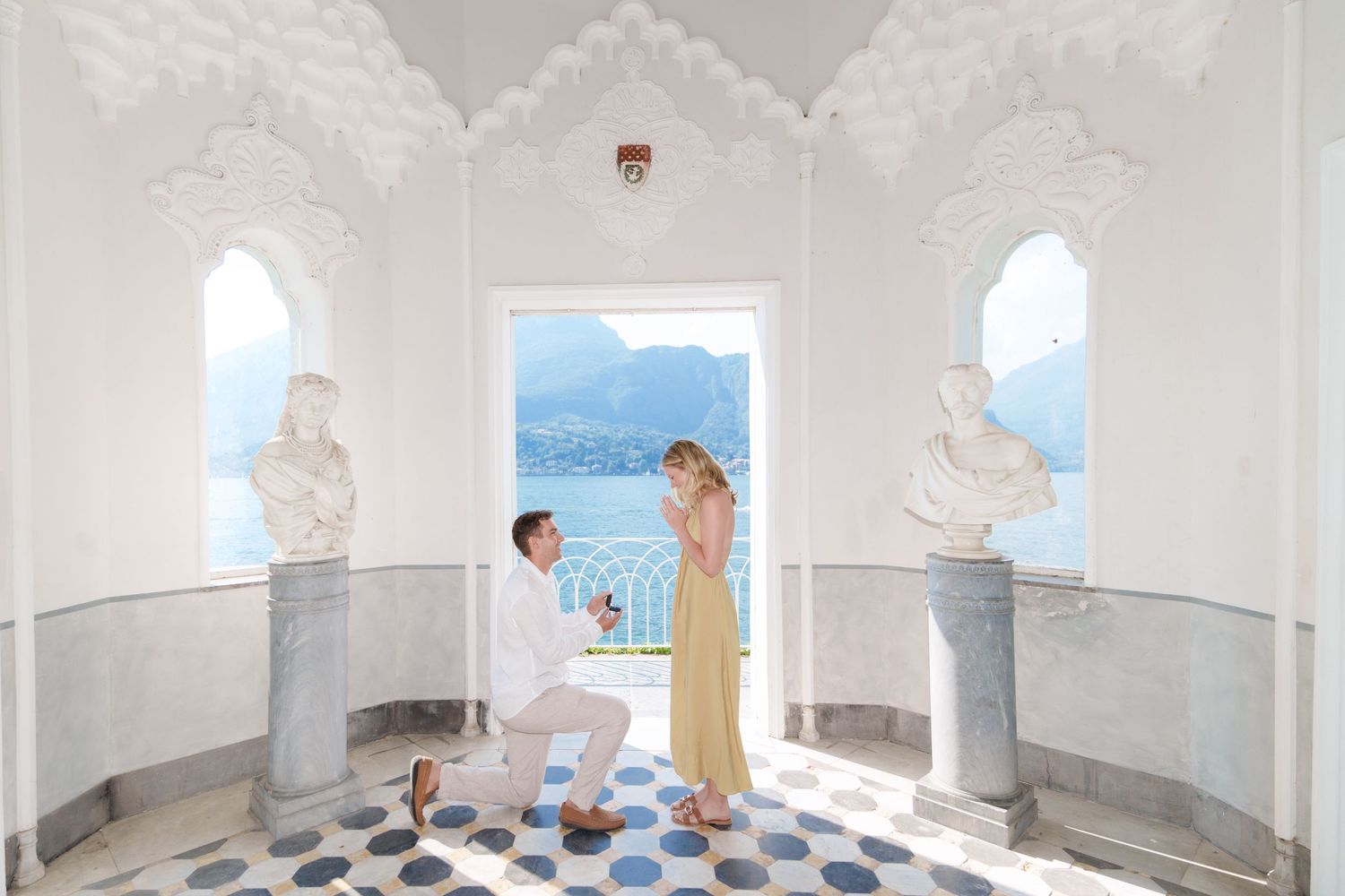 Man proposing to woman in elegant white marble hall with lake and mountain views in background.