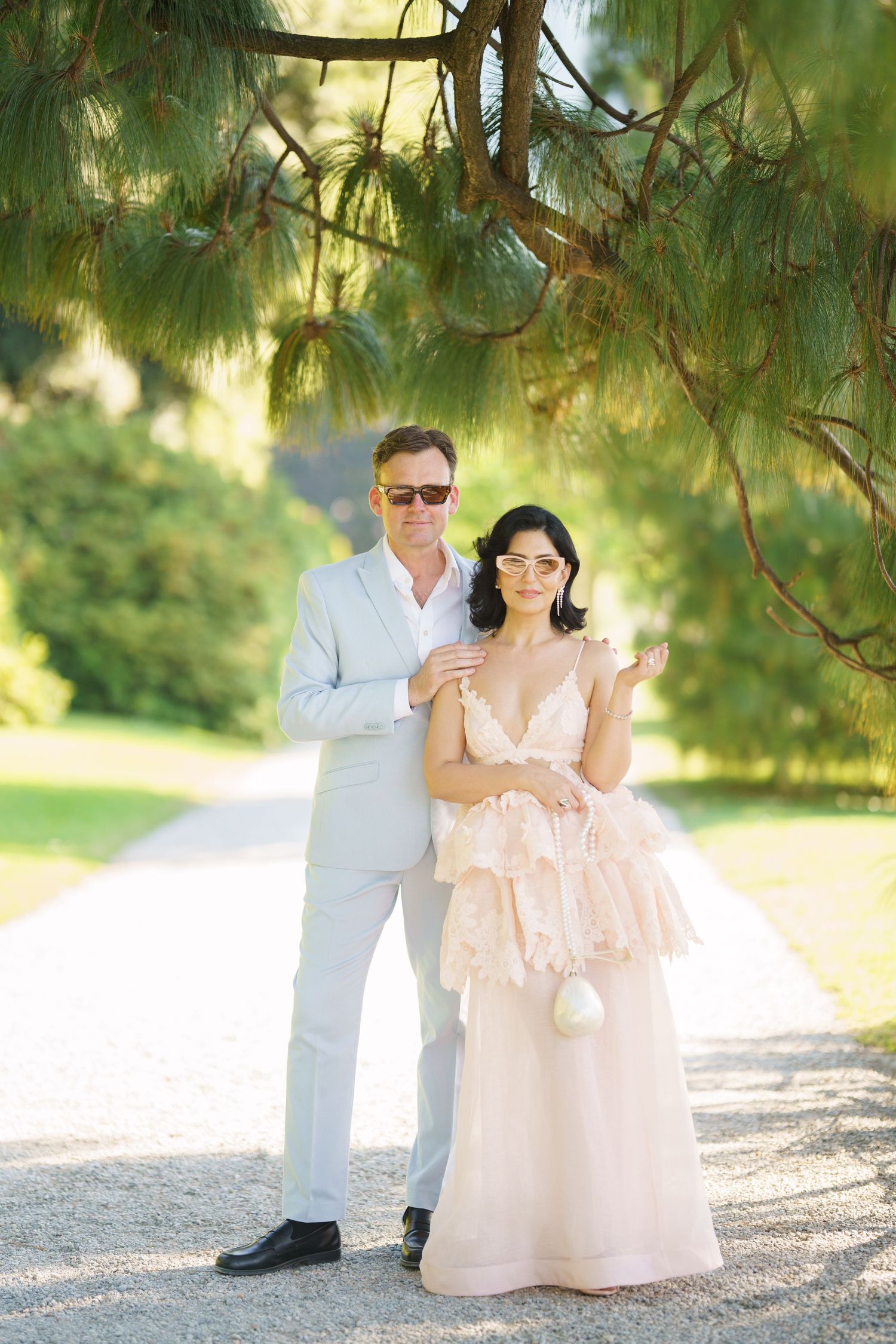 Couple posing outdoors under pine tree, man in light gray suit, woman in blush ruffled gown, garden path background.