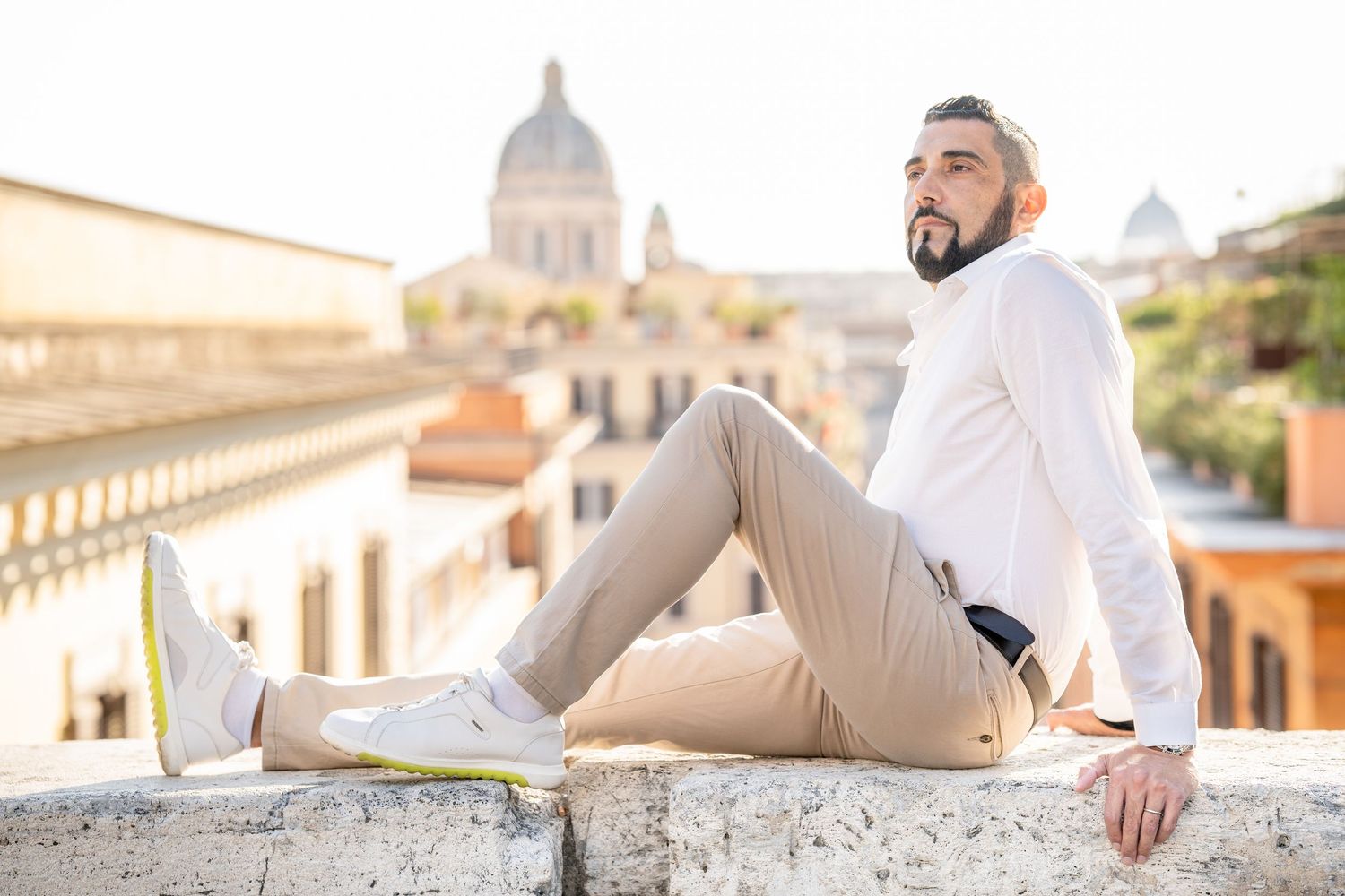 A stylish man in white shirt and beige pants sits on a stone ledge with Rome's historic skyline behind him.