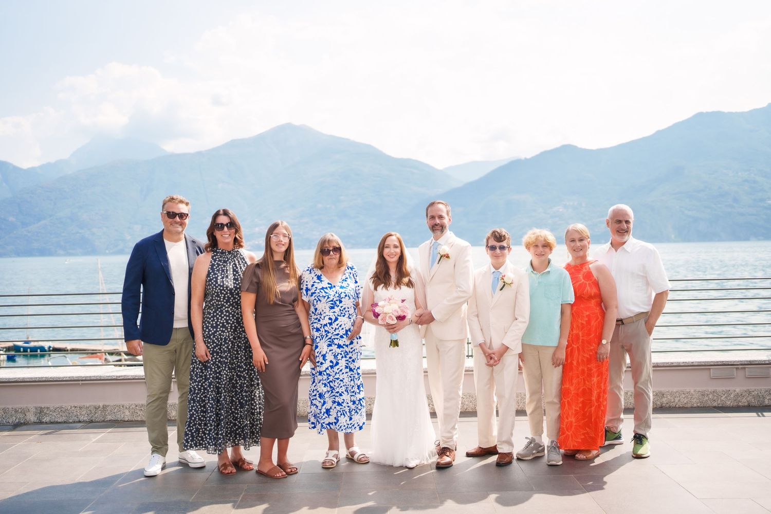 Wedding party poses by a lakeside terrace with stunning mountain backdrop on a sunny day.