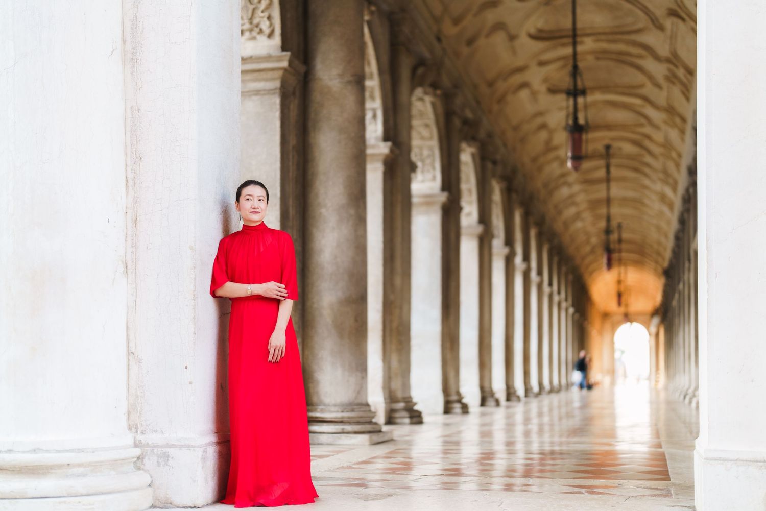 Woman in elegant red gown posing in a classical European colonnade with marble columns and arched corridor.