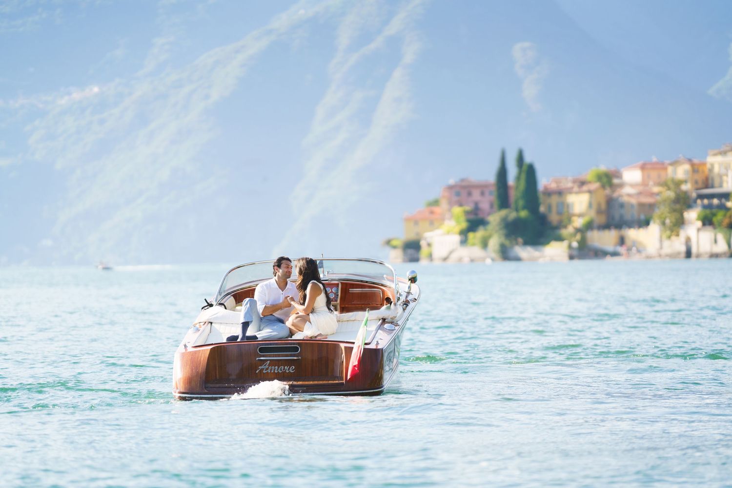 Couple enjoying a romantic boat ride on a turquoise lake with scenic mountains and a village in the background.