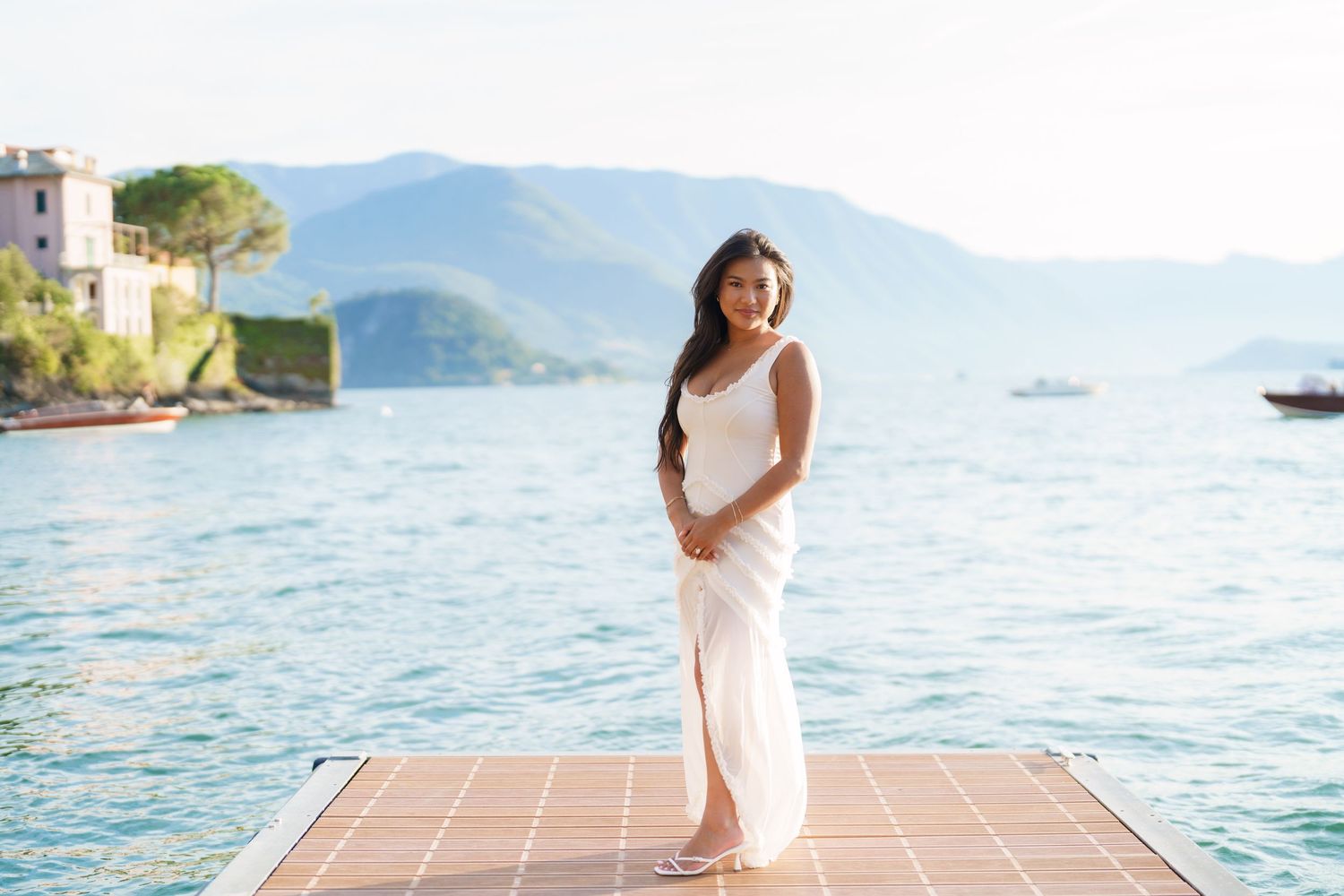 Woman in white dress standing on a dock with scenic blue lake and mountain backdrop in Italy.