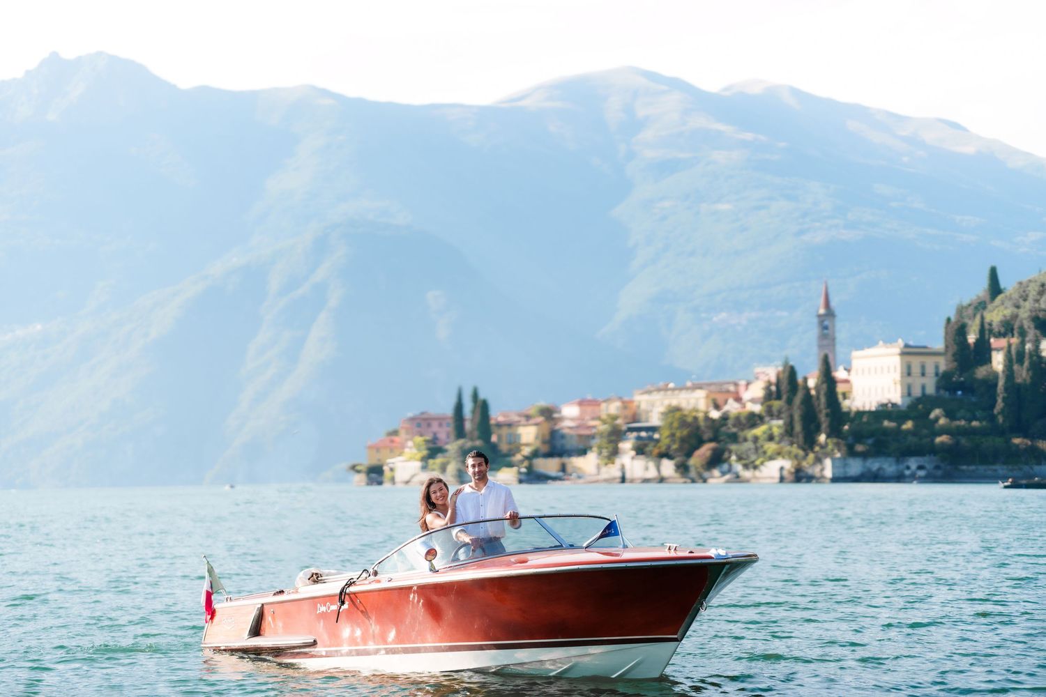 Couple riding a red vintage speedboat on Lake Como with Italian village and mountains in background.