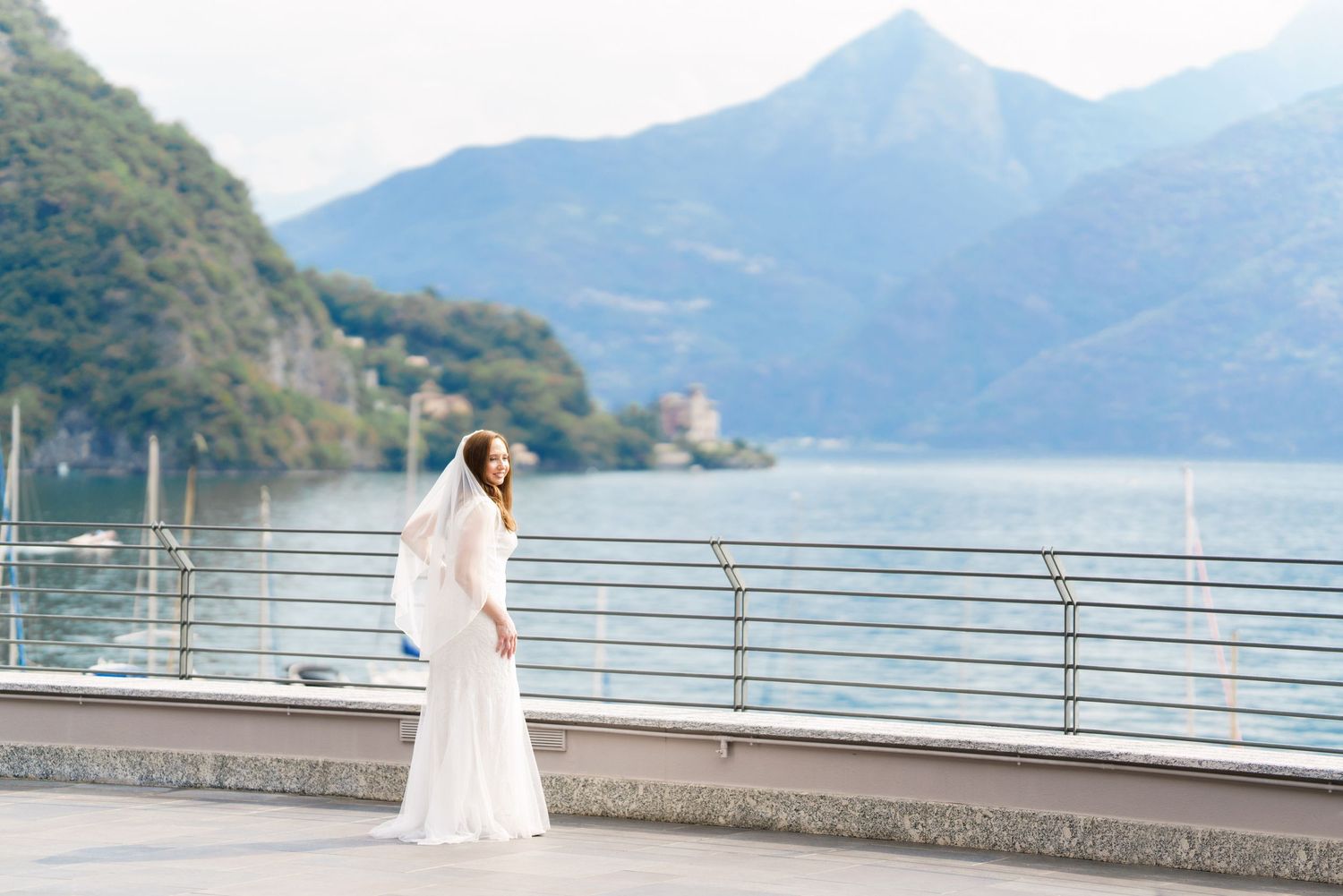 Bride in white gown and veil stands on lakeside terrace with mountain backdrop at Lake Como, Italy.