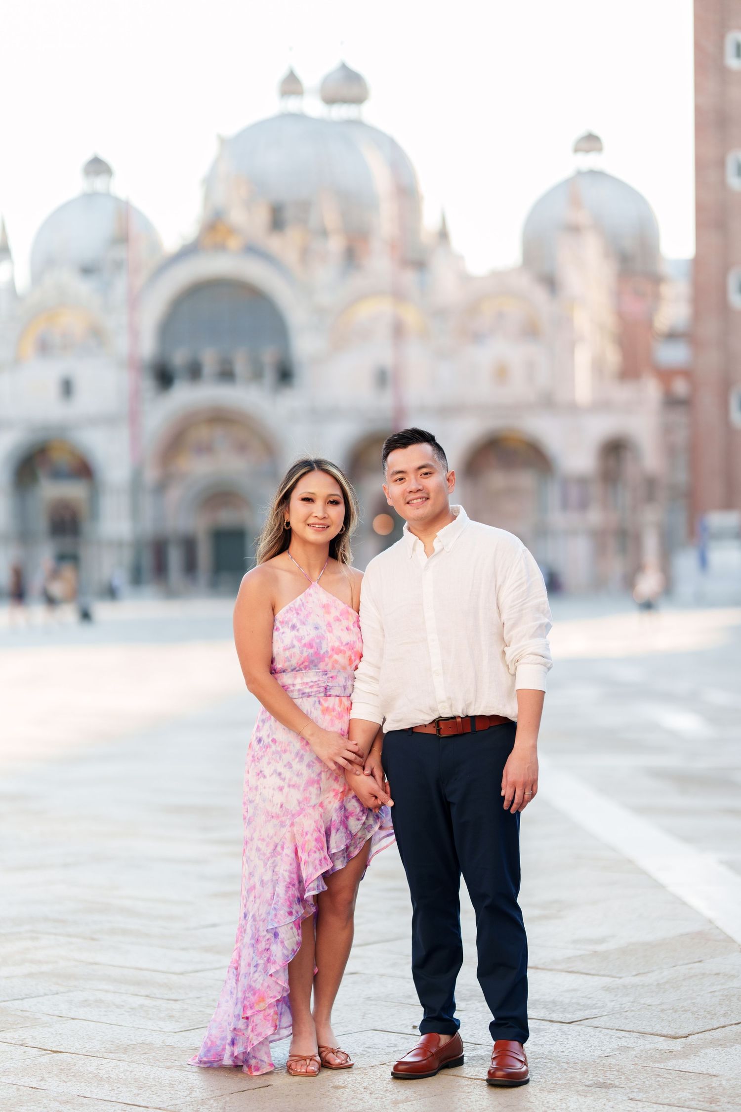 Couple posing in front of St. Mark's Basilica in Venice, woman in pink floral dress, man in white shirt.