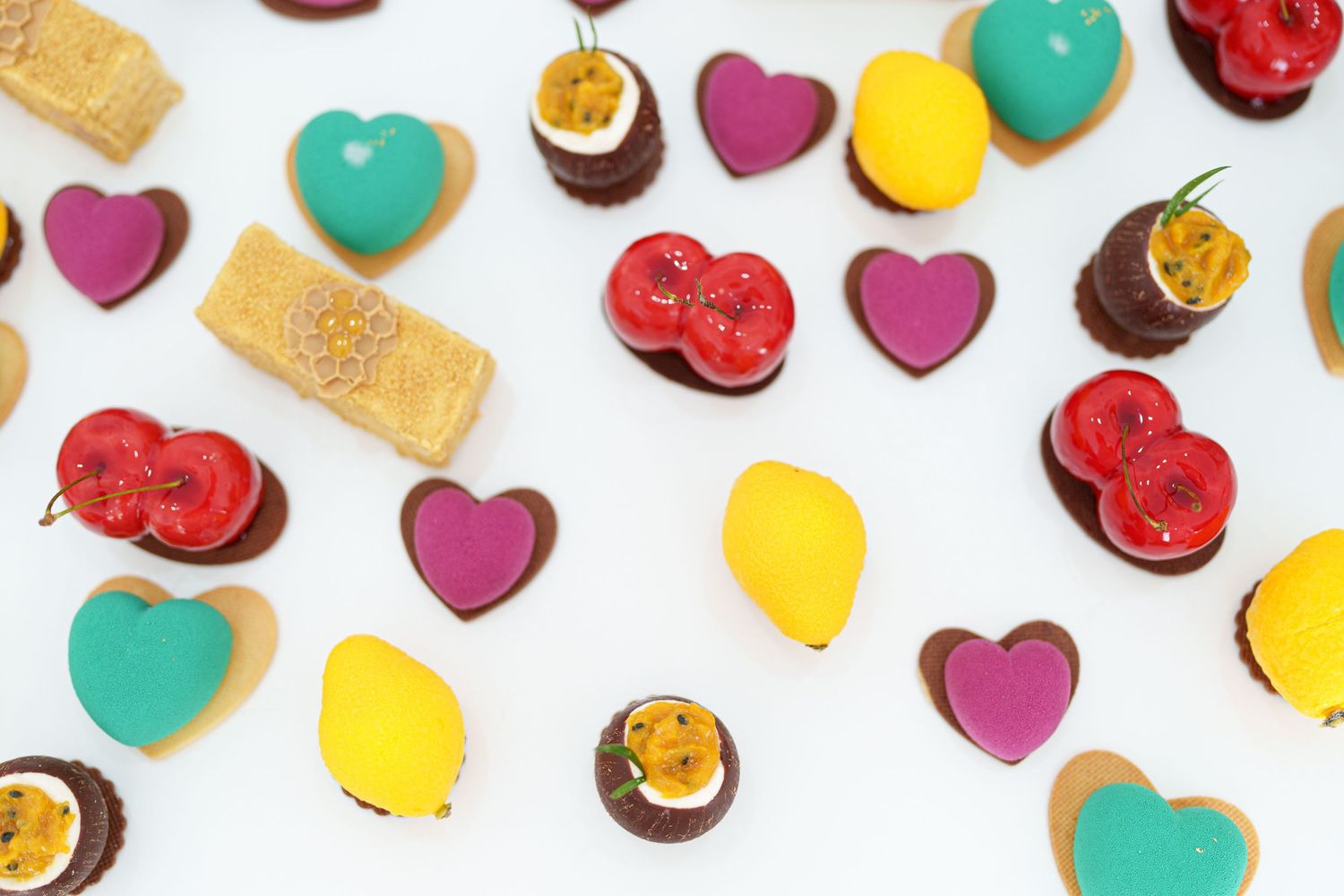 Colorful heart-shaped chocolates, macarons, and fruit-shaped sweets arranged on a white background.