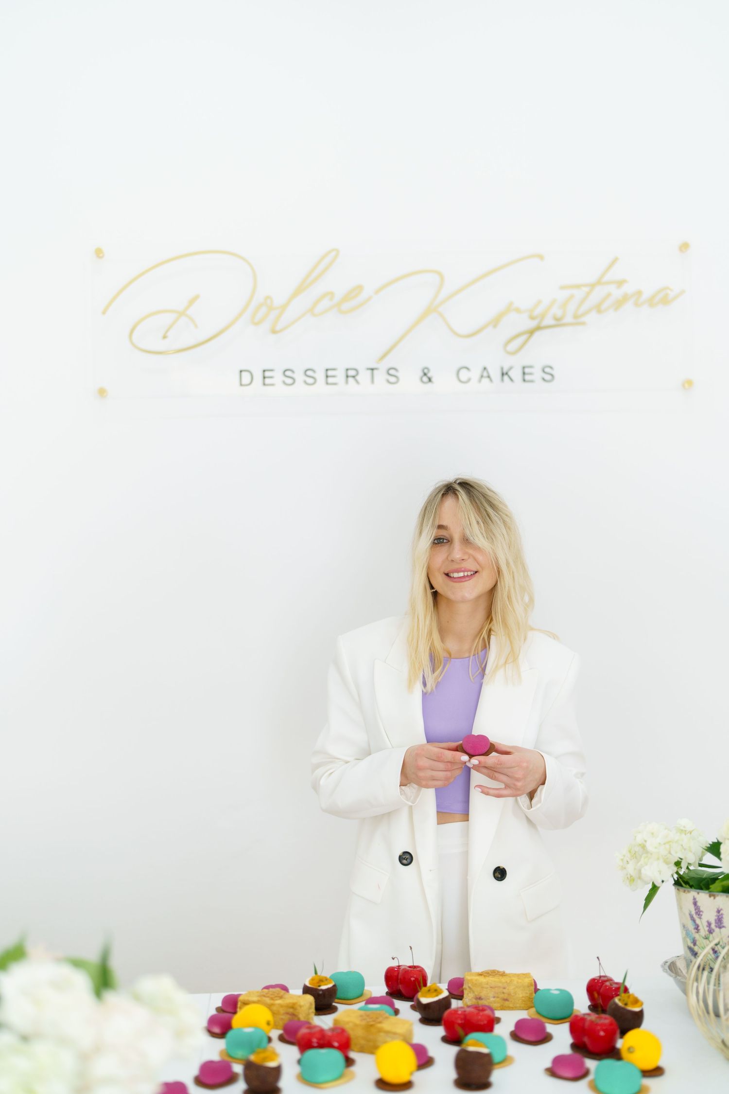 Woman in white blazer smiling behind colorful desserts at Dolce Krystina Desserts & Cakes bakery.