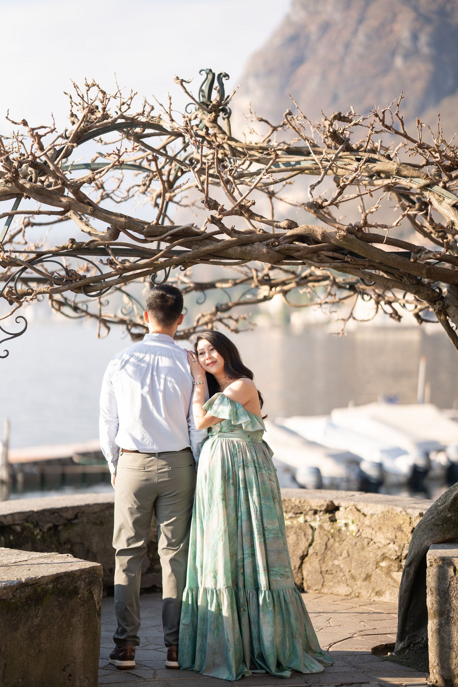 Couple in elegant attire standing under bare tree arch by lakeside with mountain backdrop.