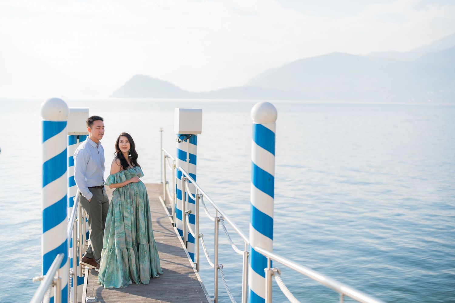 Couple posing on a blue and white striped pier overlooking calm ocean waters with misty mountains in the background.