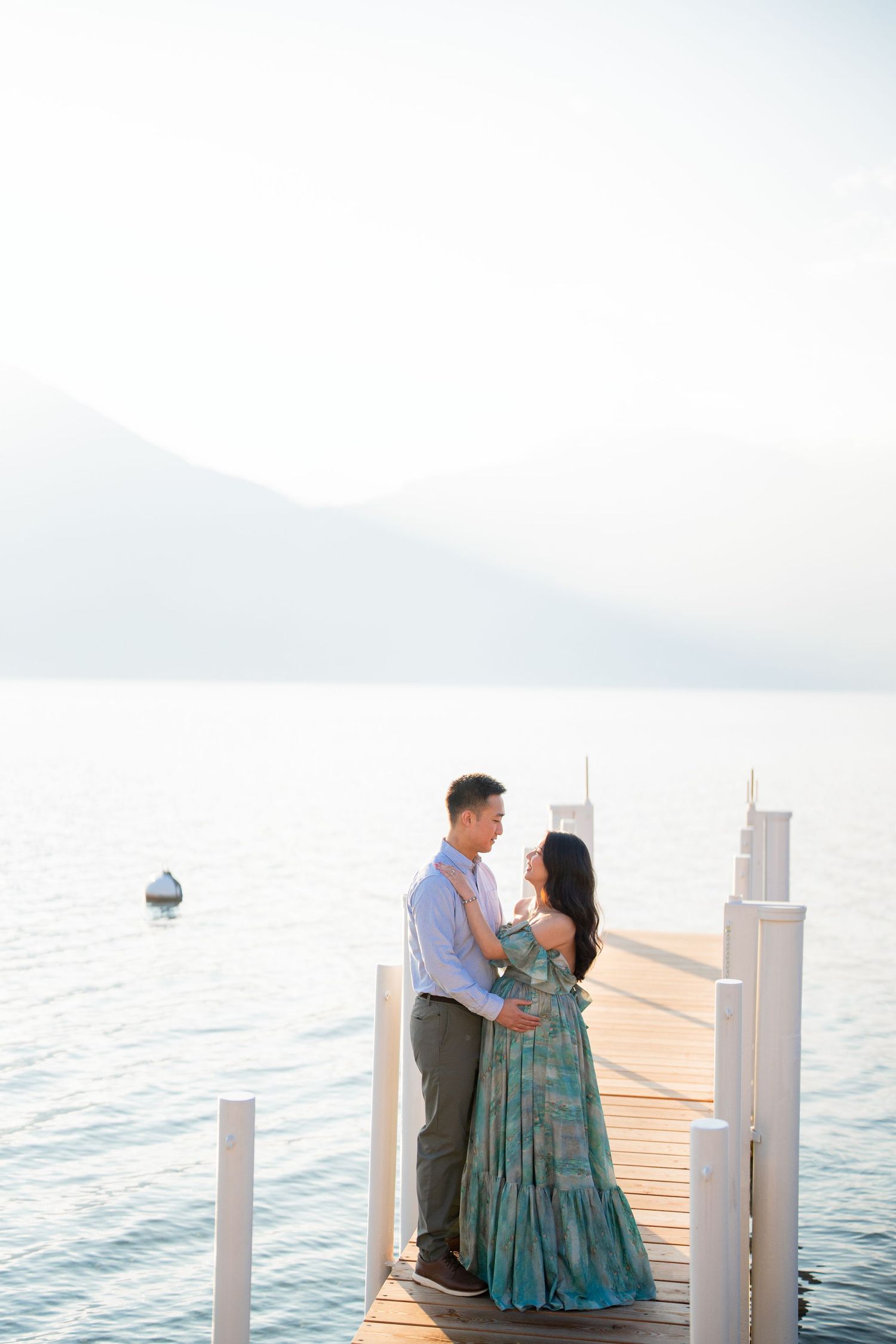 Couple embracing on a wooden pier overlooking a serene lake with misty mountains in the background.