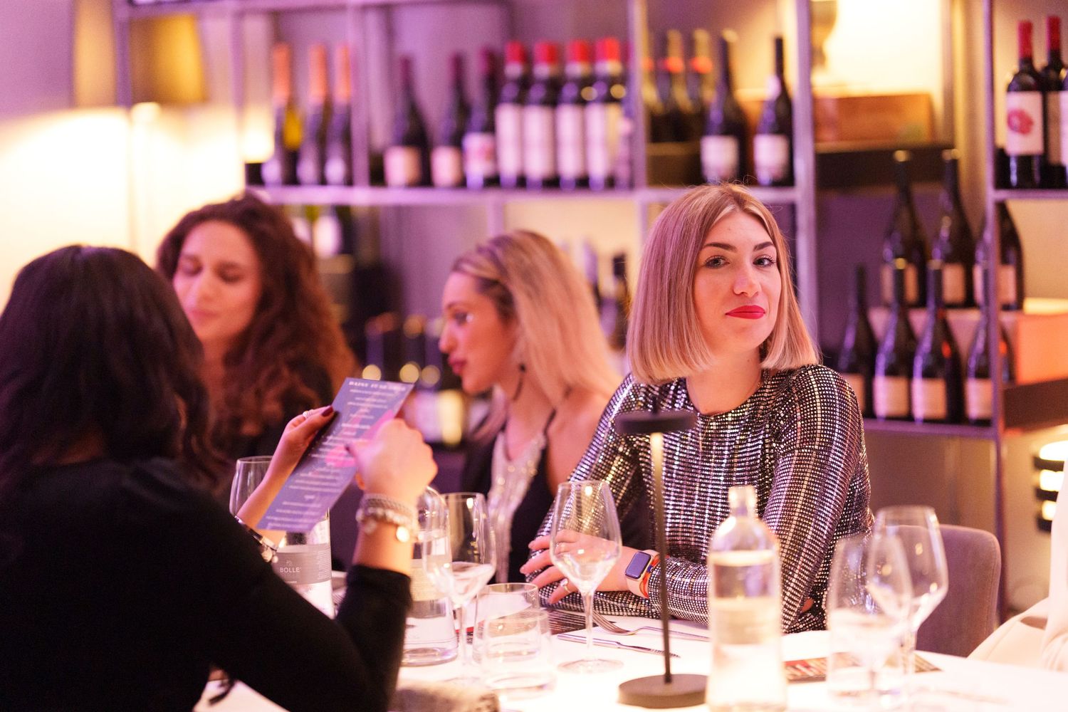 Women socializing at a wine bar dinner event, with bottles displayed on shelves in a warm, elegant setting.