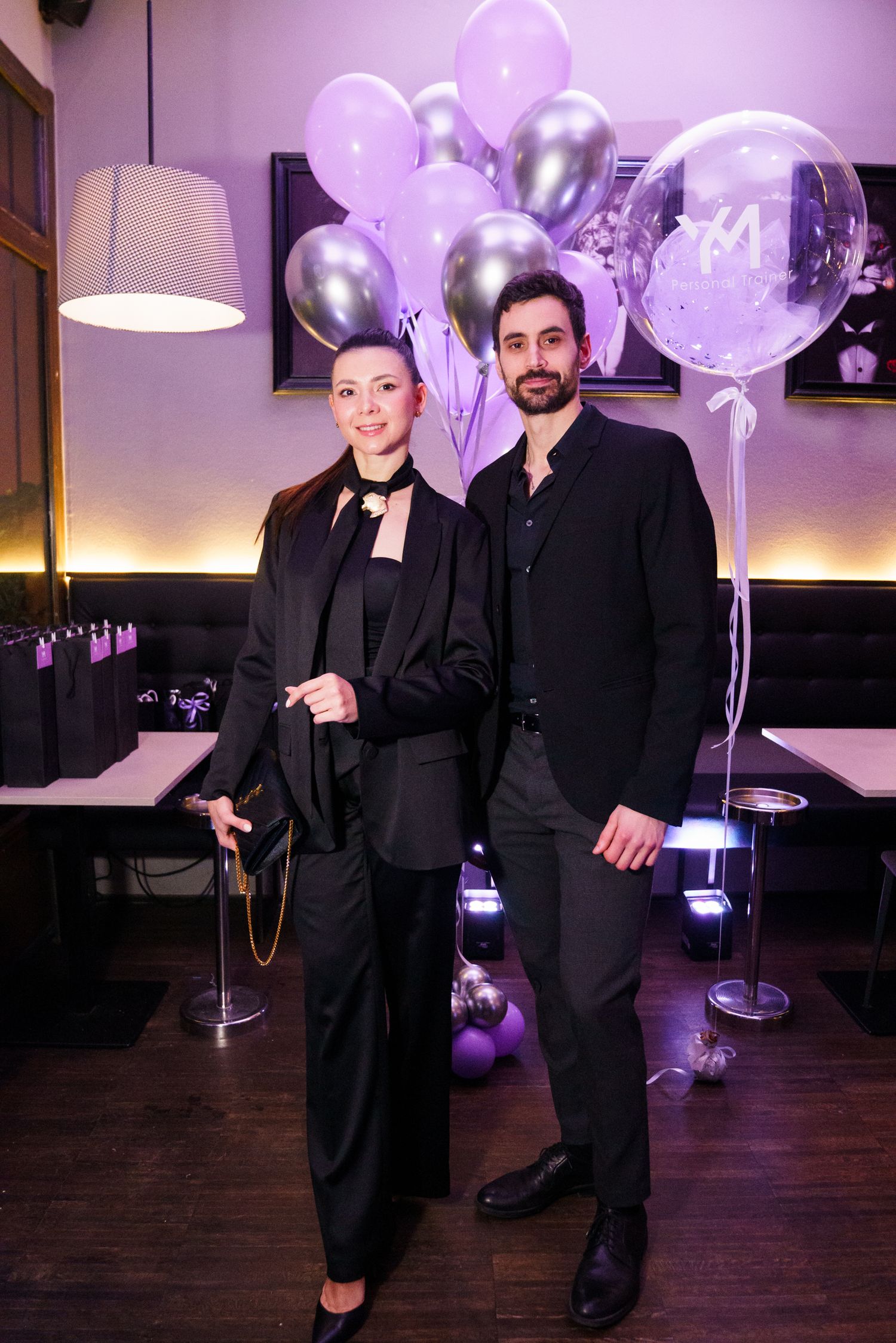 Stylish couple dressed in black posing together at a purple-themed party with metallic balloons in a decorated venue.