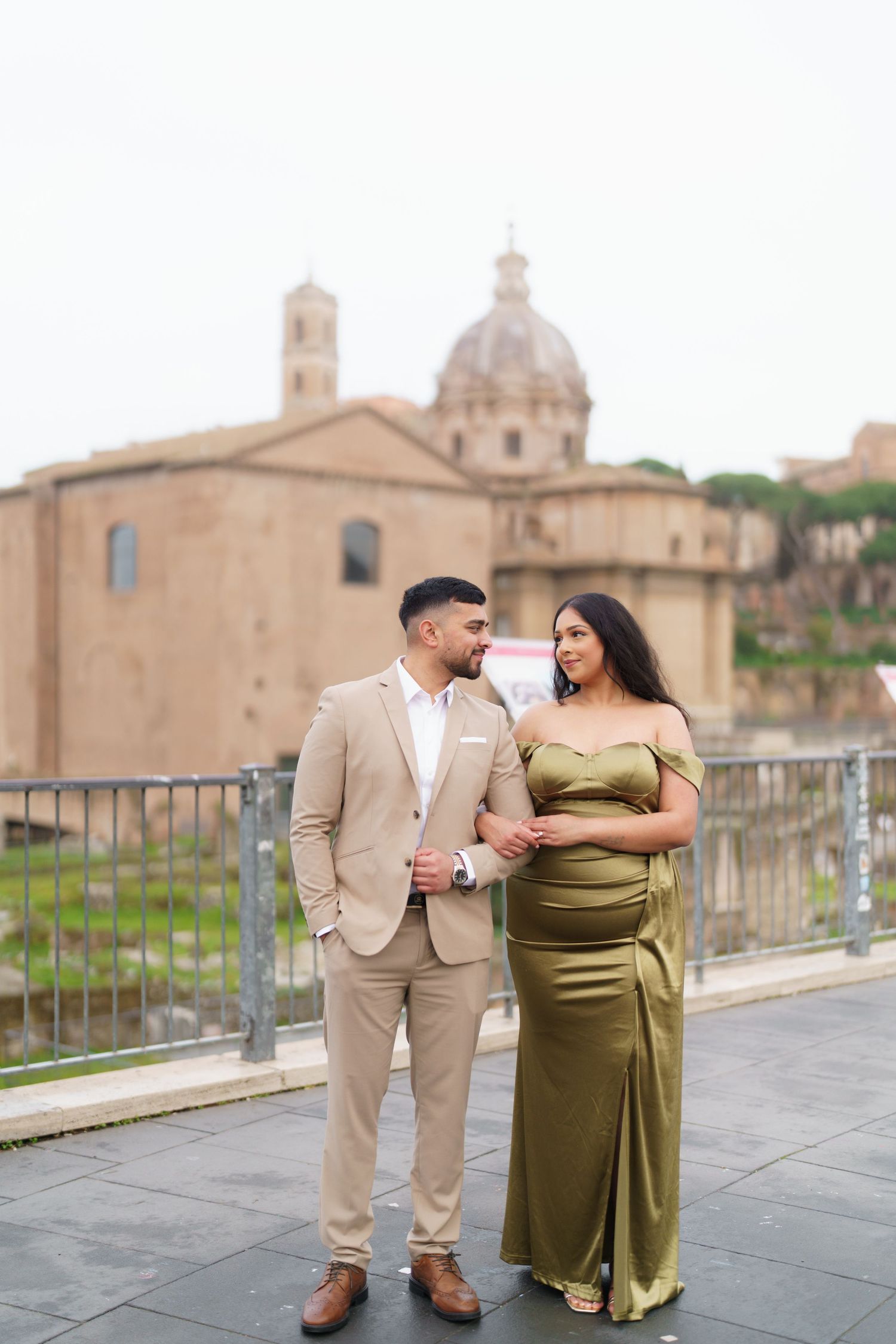 Elegant couple posing in Rome, man in beige suit and woman in olive green gown, with historic Roman architecture behind.