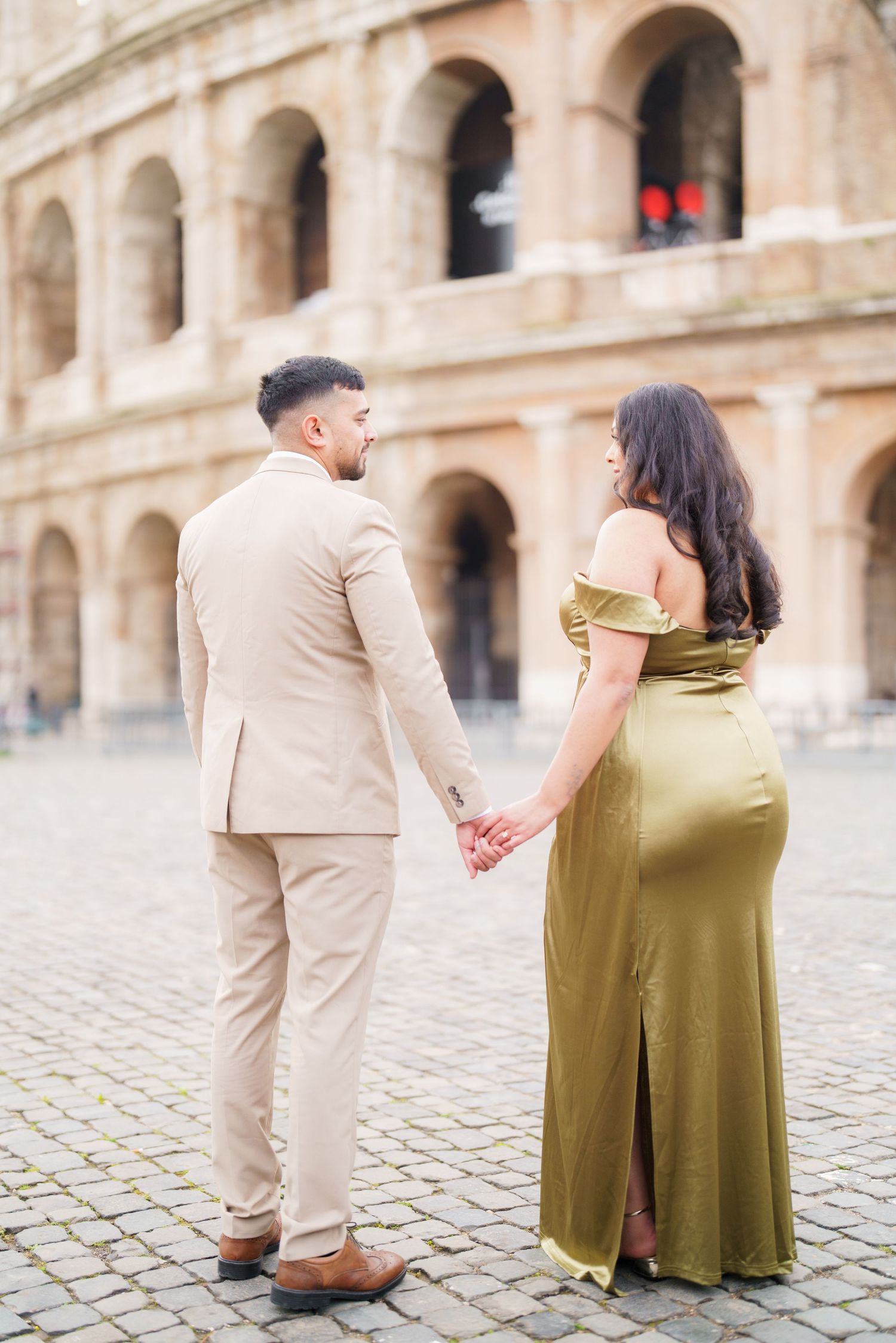 Couple holding hands in front of the Colosseum in Rome, man in beige suit, woman in olive satin gown.