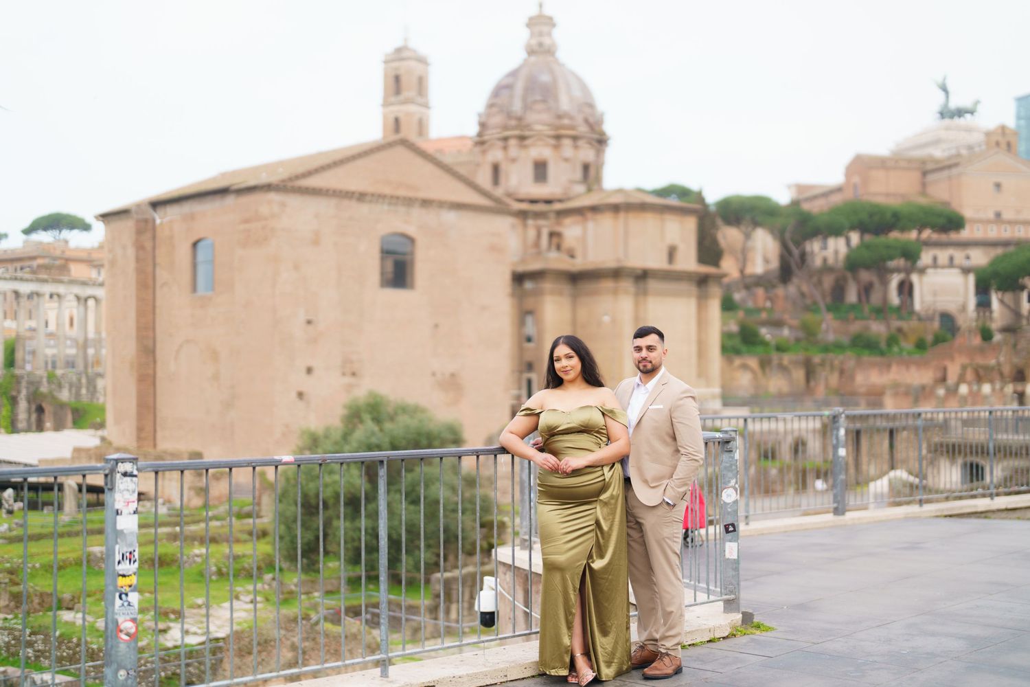 Couple posing near Roman Forum ruins in Rome, Italy, woman in gold gown, man in beige suit, ancient architecture background.