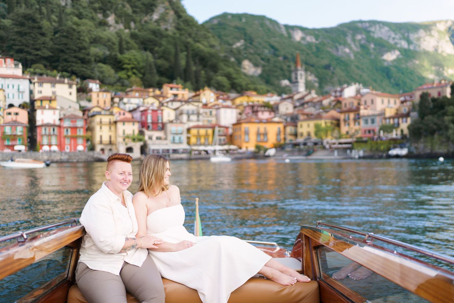 Couple sitting together on a wooden boat with colorful Italian village and green hills of Lake Como in background.
