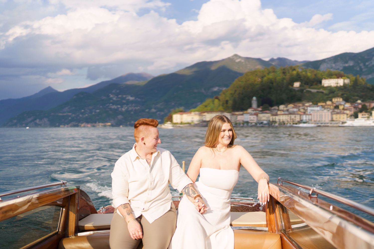 Couple holding hands on a wooden boat on Lake Como, Italy, with mountains and Bellagio village in the background.