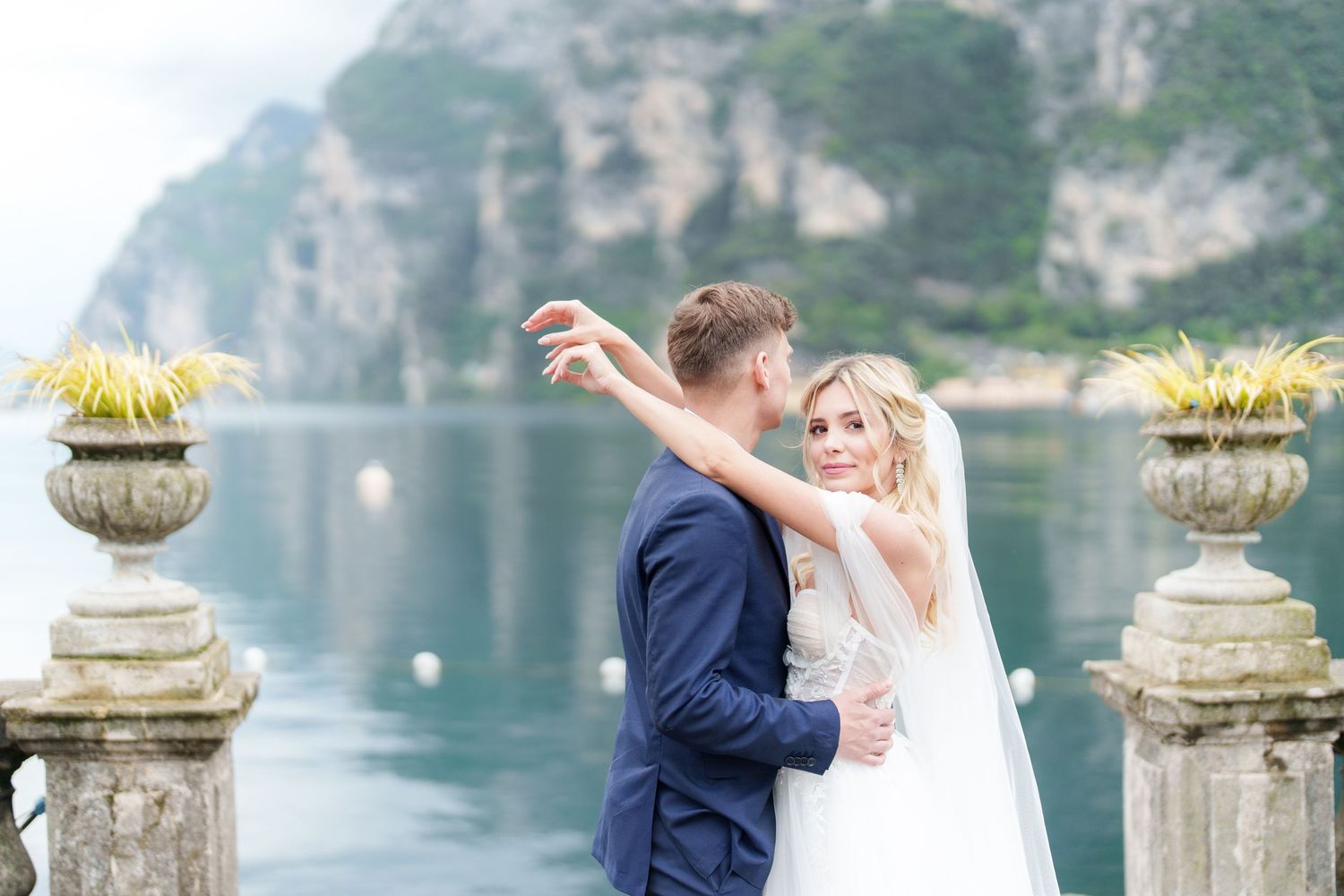 Couple embracing on elegant lakeside terrace with mountain backdrop on their wedding day.