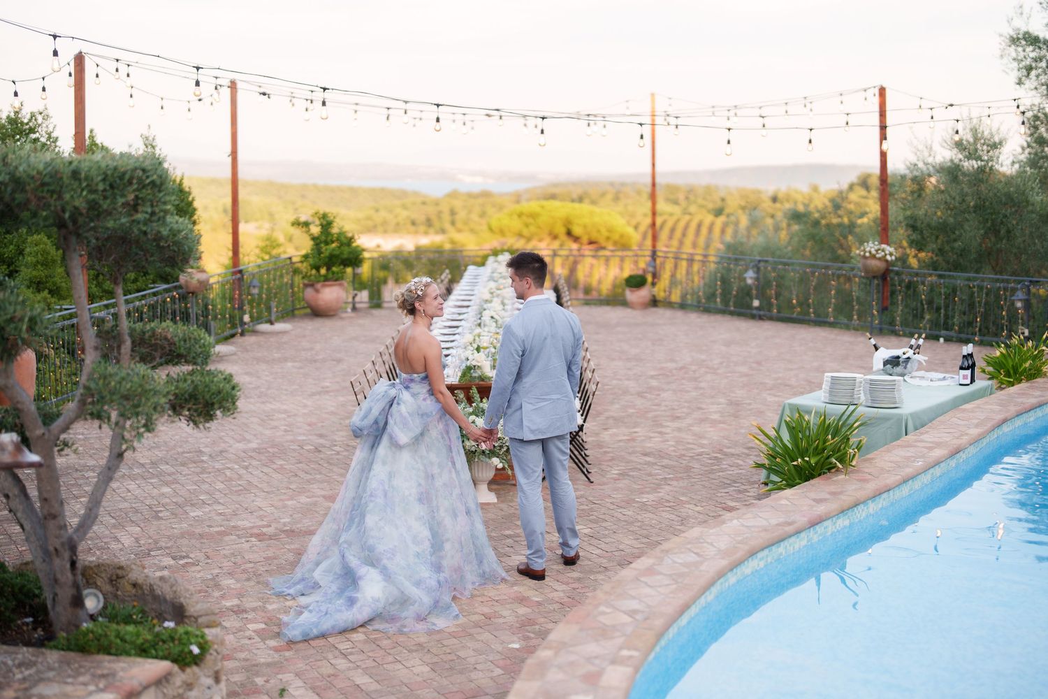 Bride and groom walk hand-in-hand beside a pool at a vineyard wedding venue during golden hour.
