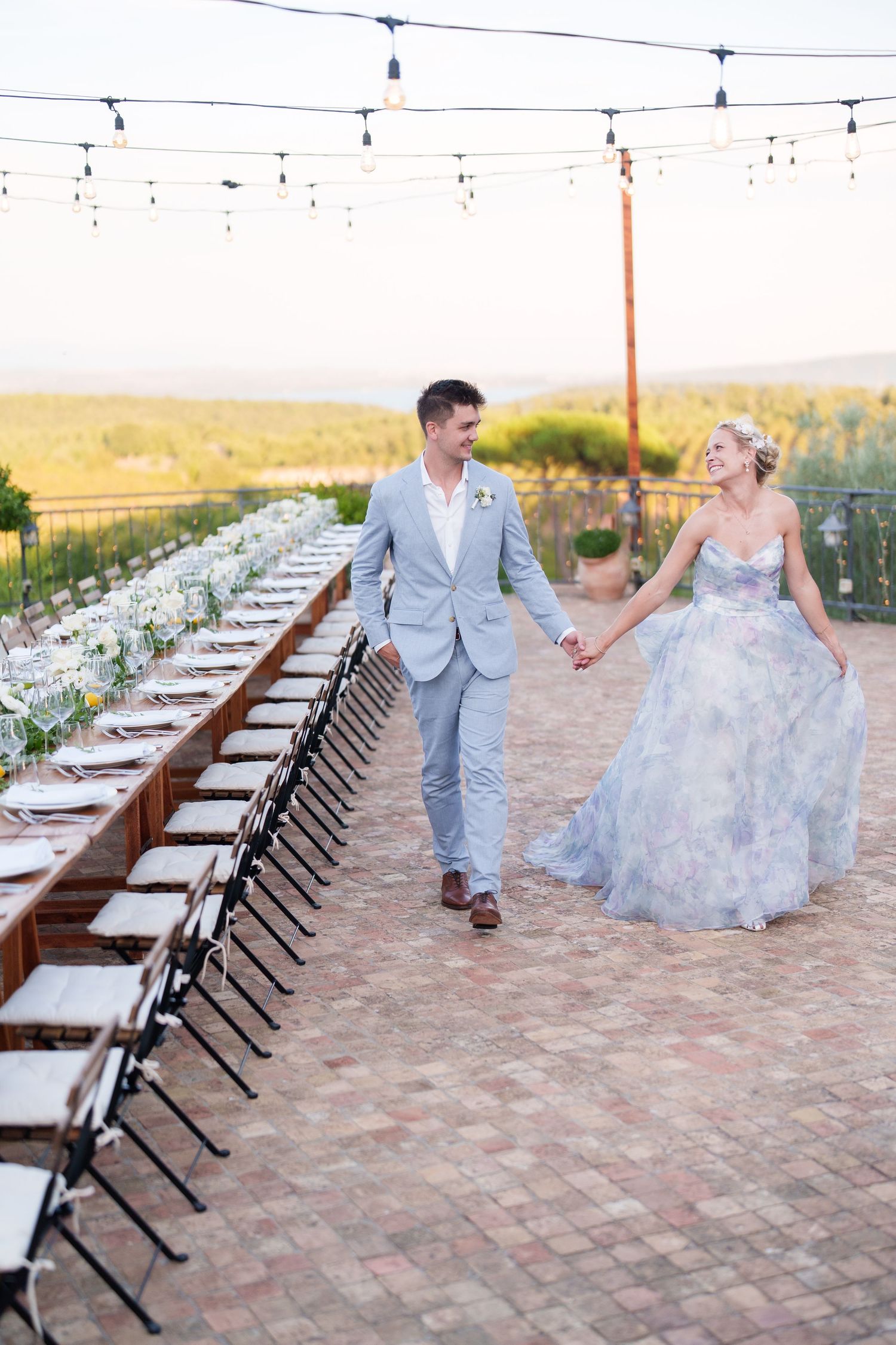 Bride in floral gown and groom in gray suit walk hand-in-hand along long reception table on brick patio at sunset.
