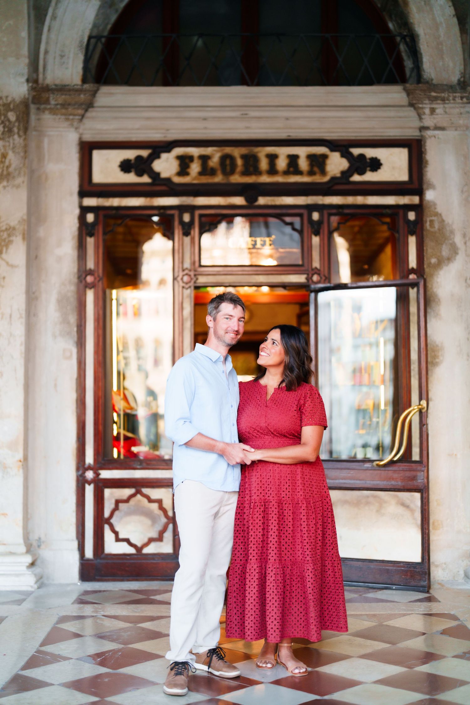Couple smiling and holding hands in front of Cafe Florian in Venice, Italy, woman in red dress.