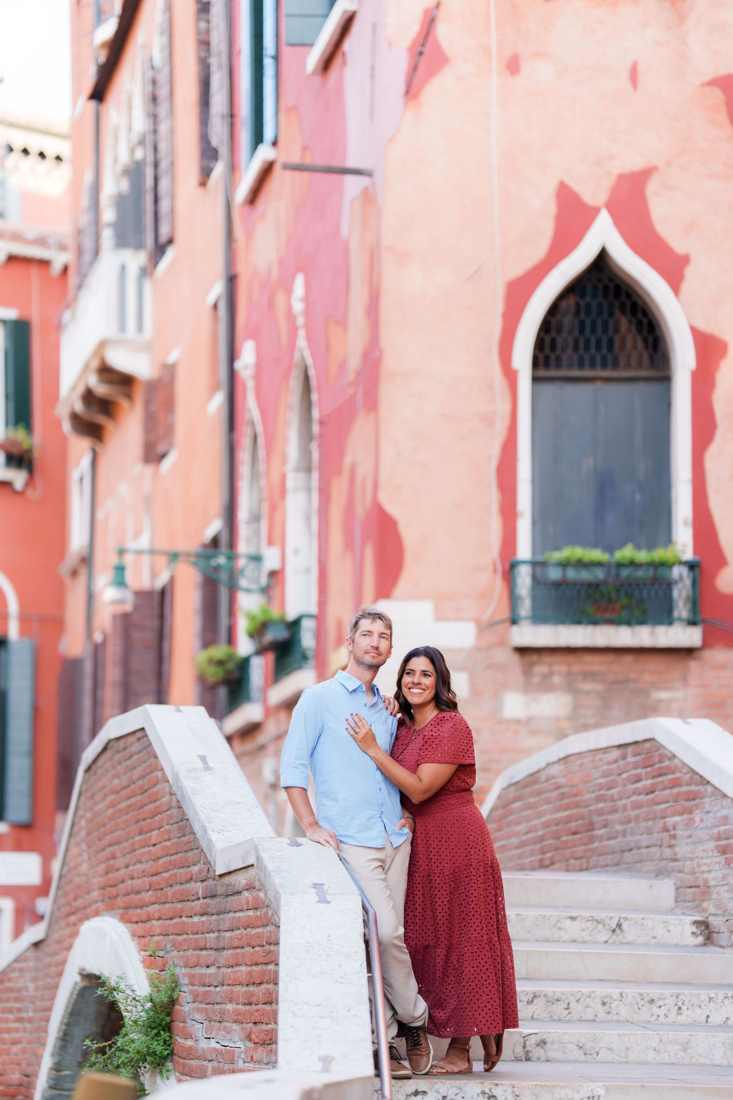 Couple posing on a bridge in Venice, Italy, with colorful pink and red historic buildings in the background.