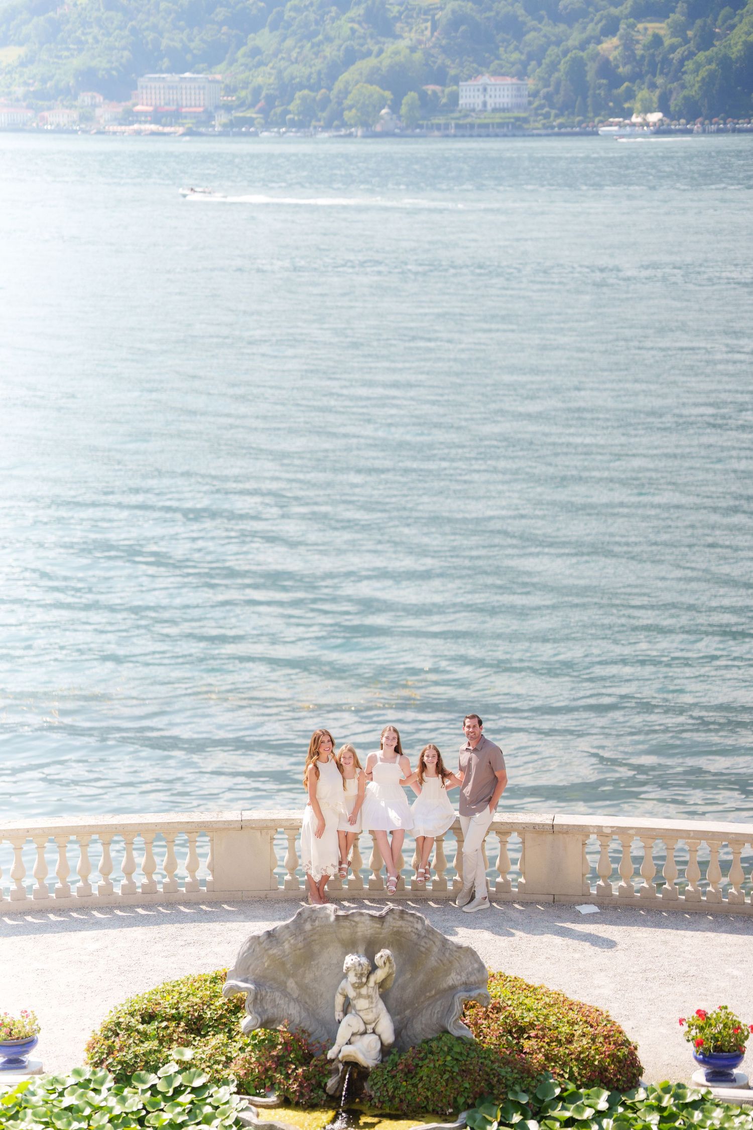 Family in white outfits posing on elegant lakeside terrace with ornate balustrade, lush greenery, and scenic blue water backdrop.