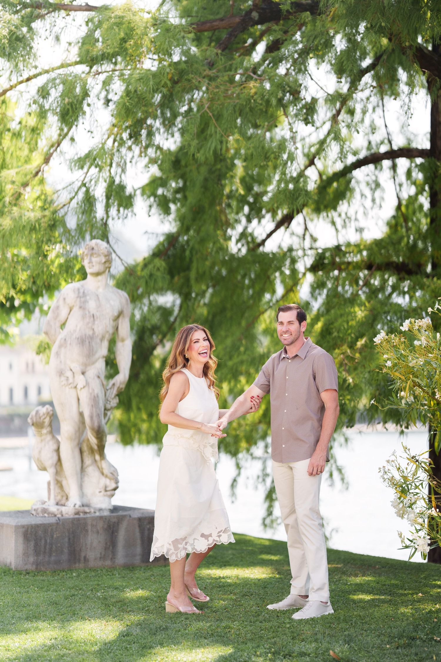 Couple holding hands and laughing near a classical statue in a lush garden by the waterfront.